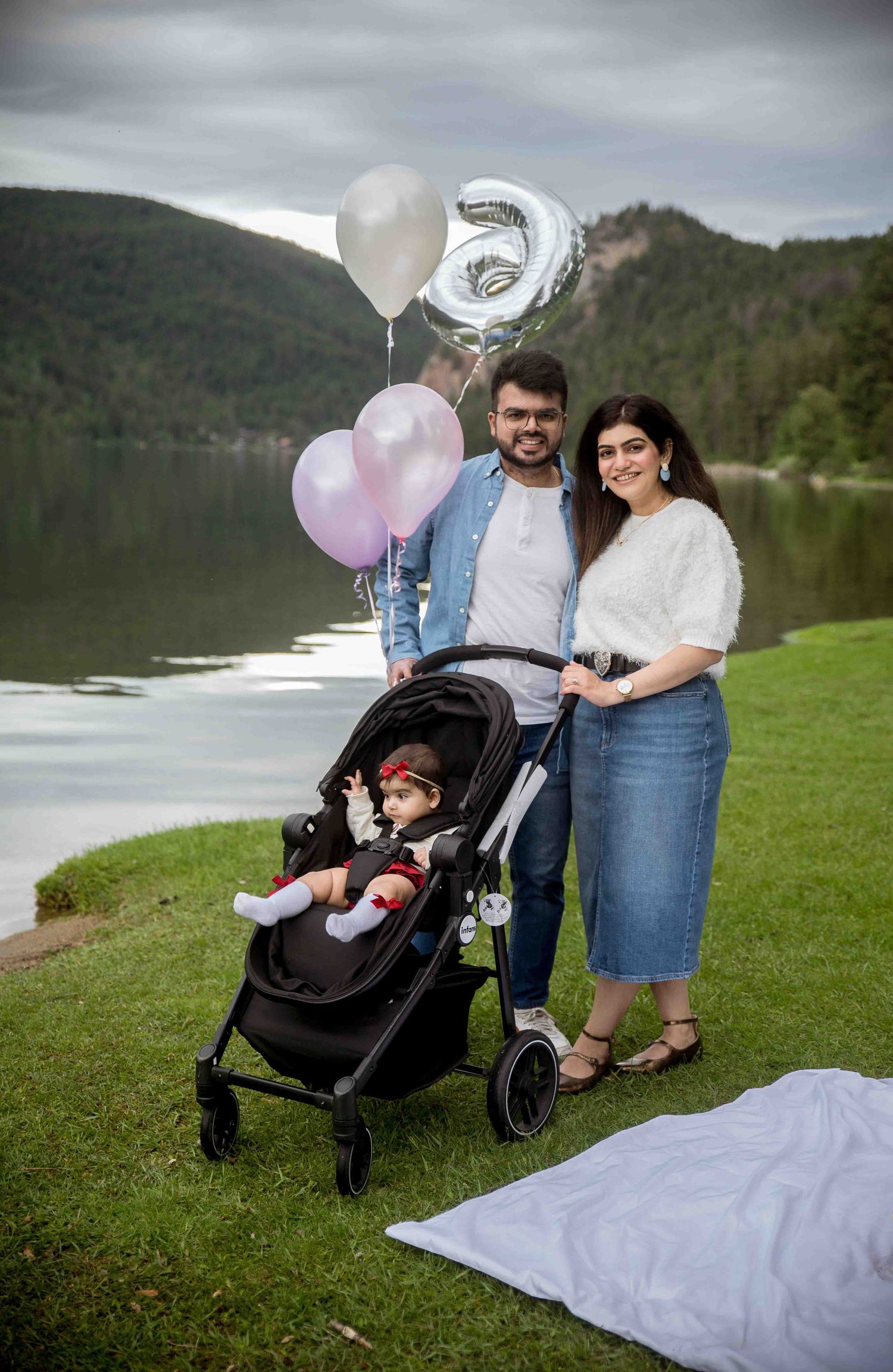 Family posing with balloons and stroller by a lake for a baby's 6-month celebration outdoors.