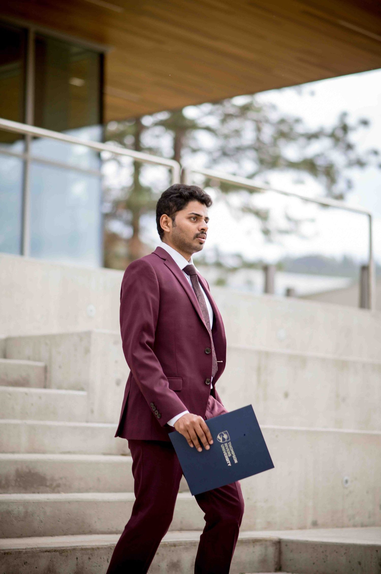 Graduate in maroon suit carrying a diploma folder down outdoor concrete steps.