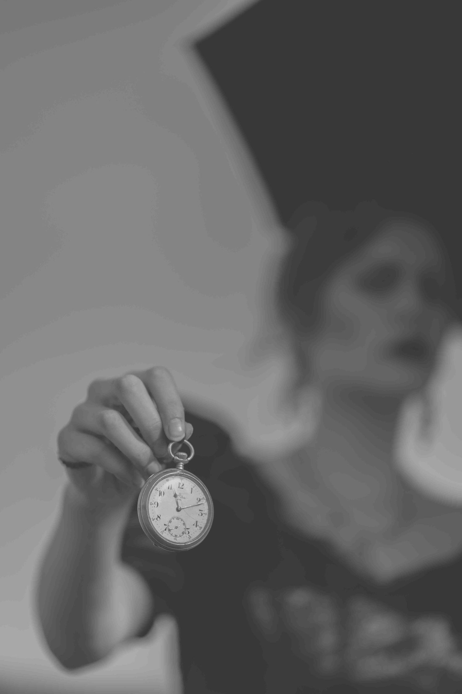 Black and white photo of a woman holding a vintage pocket watch toward the camera, with a blurred figure in the background.