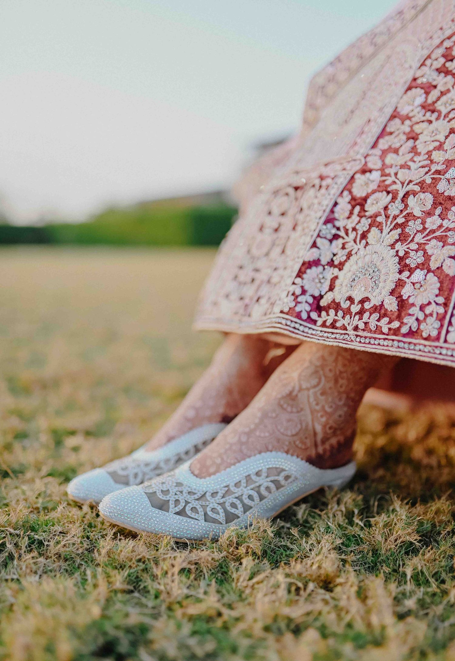 Close-up of bridal mehndi henna feet wearing white embroidered juttis with red embroidered lehenga on grass.