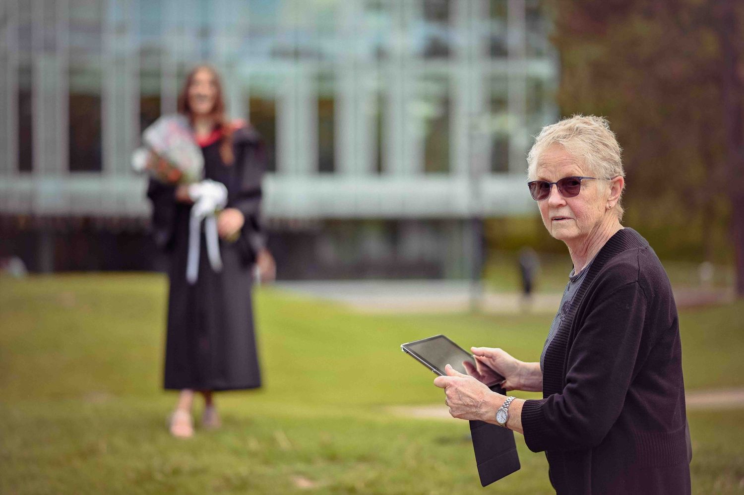 Older woman holding tablet outdoors with graduate in black gown standing in background near glass building.