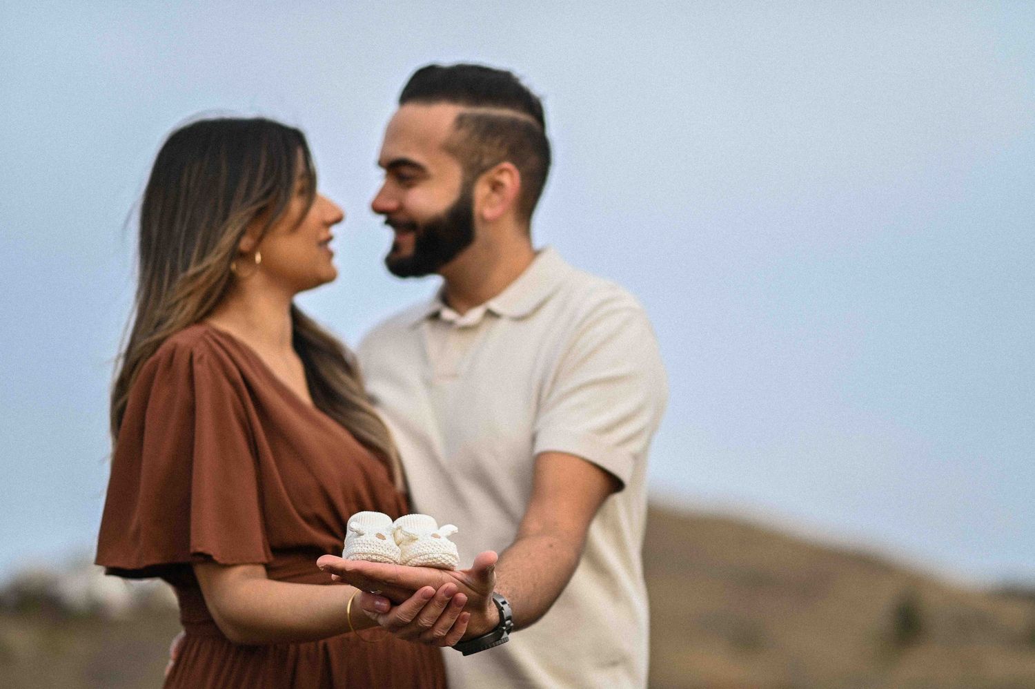 Expecting couple facing each other outdoors, holding tiny baby shoes together against a scenic backdrop.