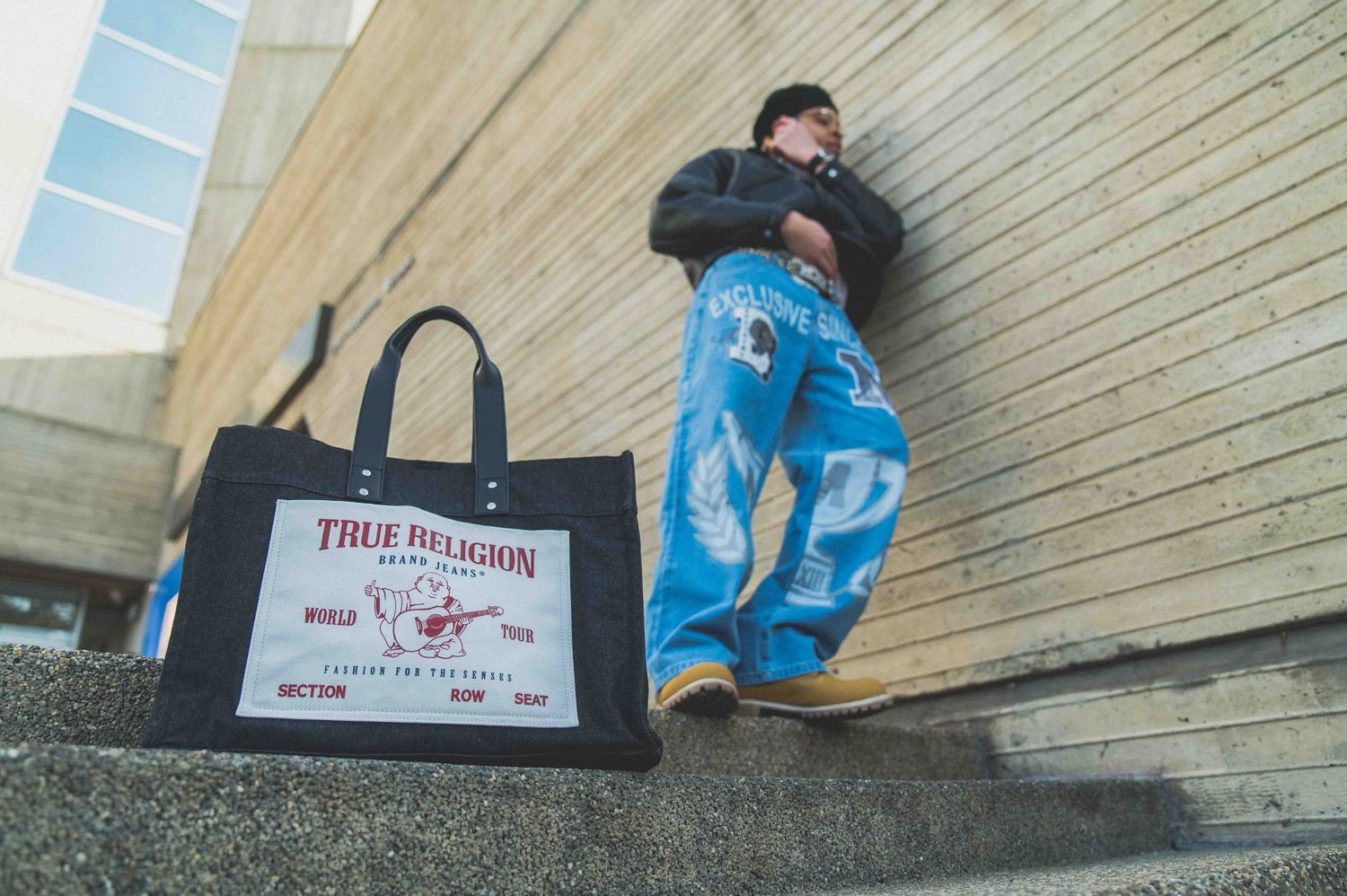 Stylish man leaning against wall wearing True Religion tote bag and blue printed jeans on urban steps.
