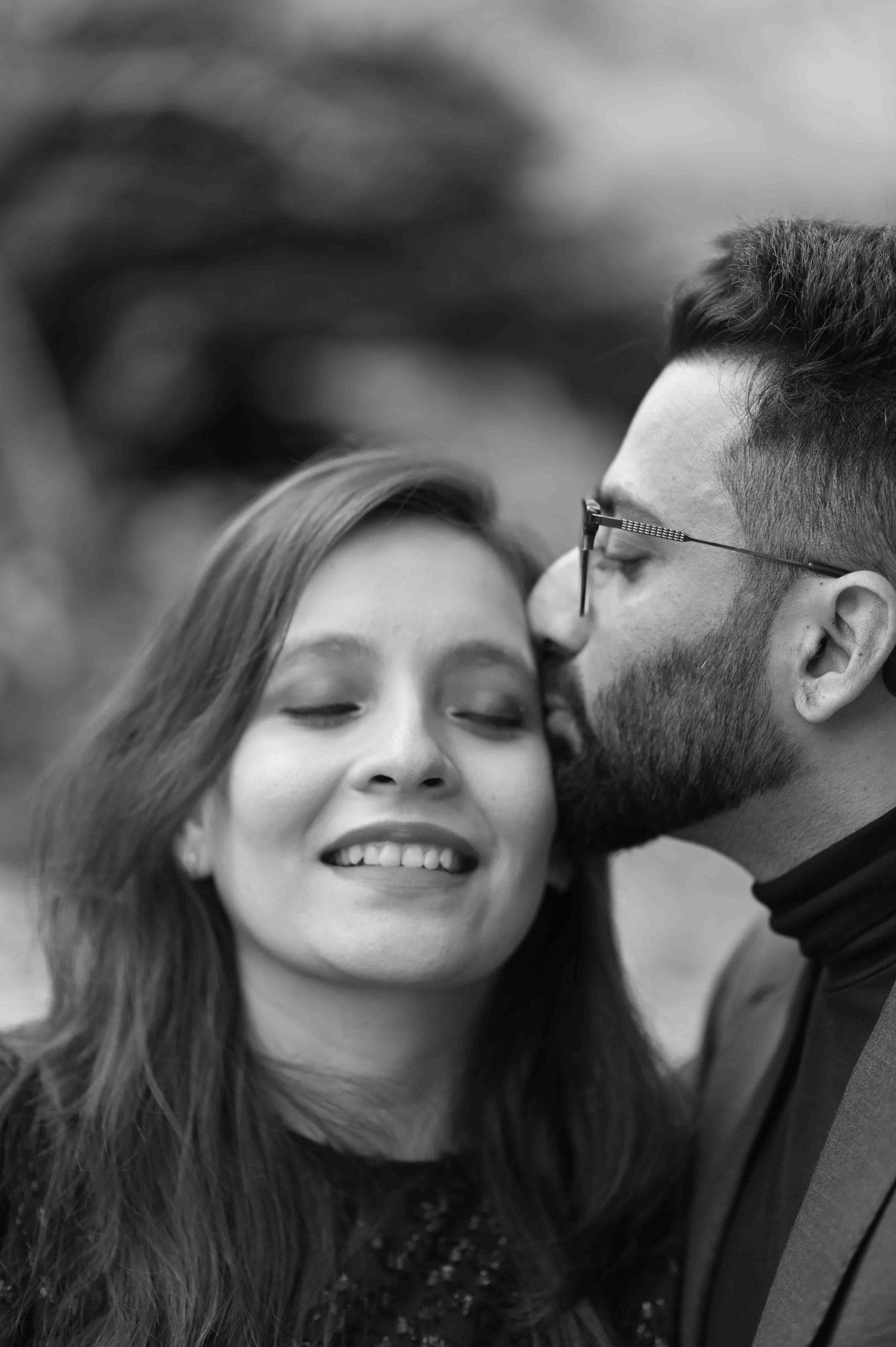 Black and white photo of a bearded man kissing a smiling woman on the cheek outdoors.