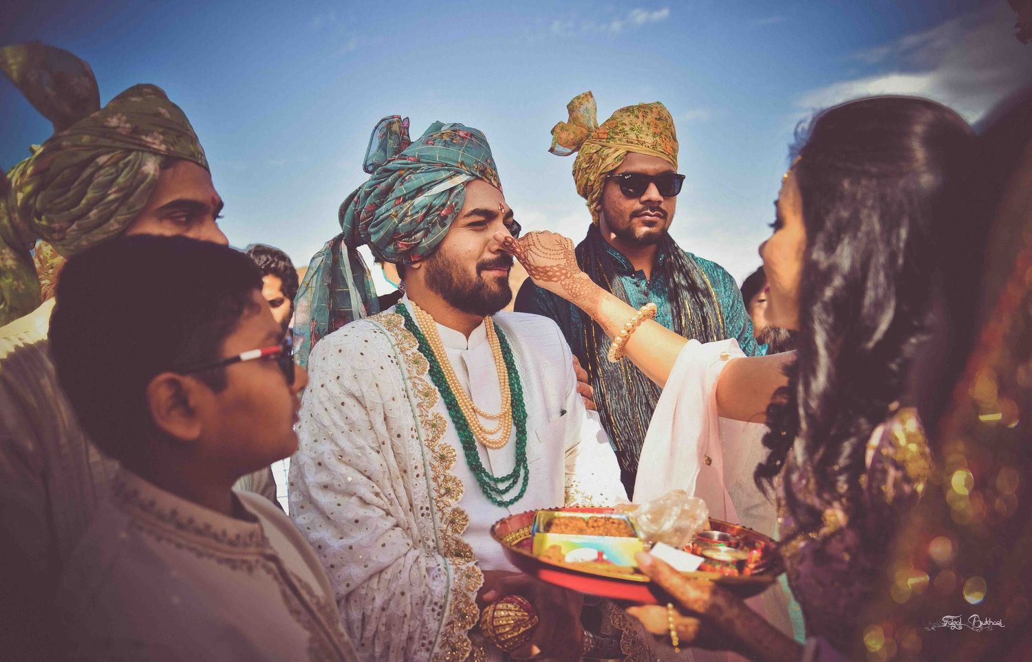 Groom in white sherwani and teal turban receives blessings during colorful outdoor Indian wedding celebration.