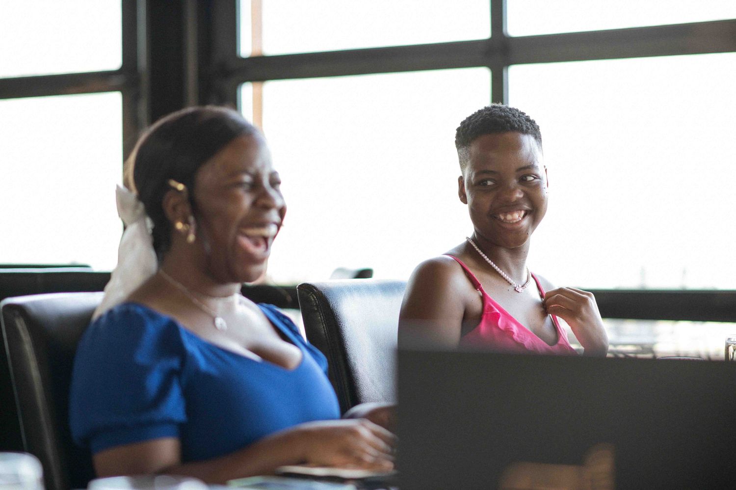 Two Black women smiling together on a couch, one in blue dress using laptop, one in pink top.