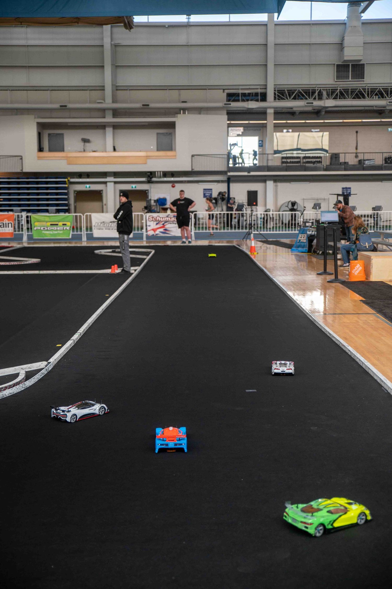 RC cars race on an indoor black track with two people standing near the course in a large sports facility.