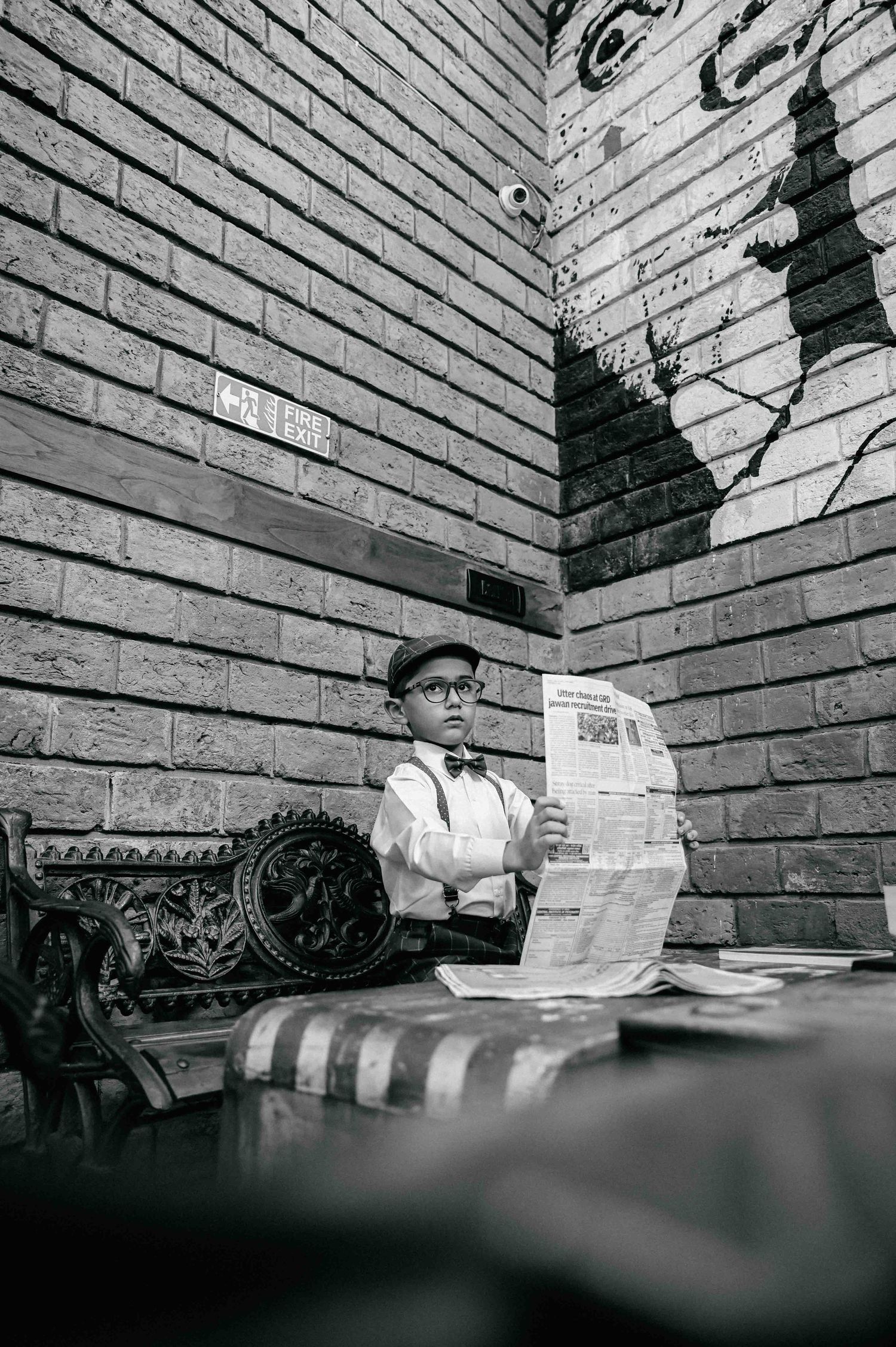 A dapper young boy in suspenders, bow tie, and newsboy cap sits reading a newspaper against a brick wall.