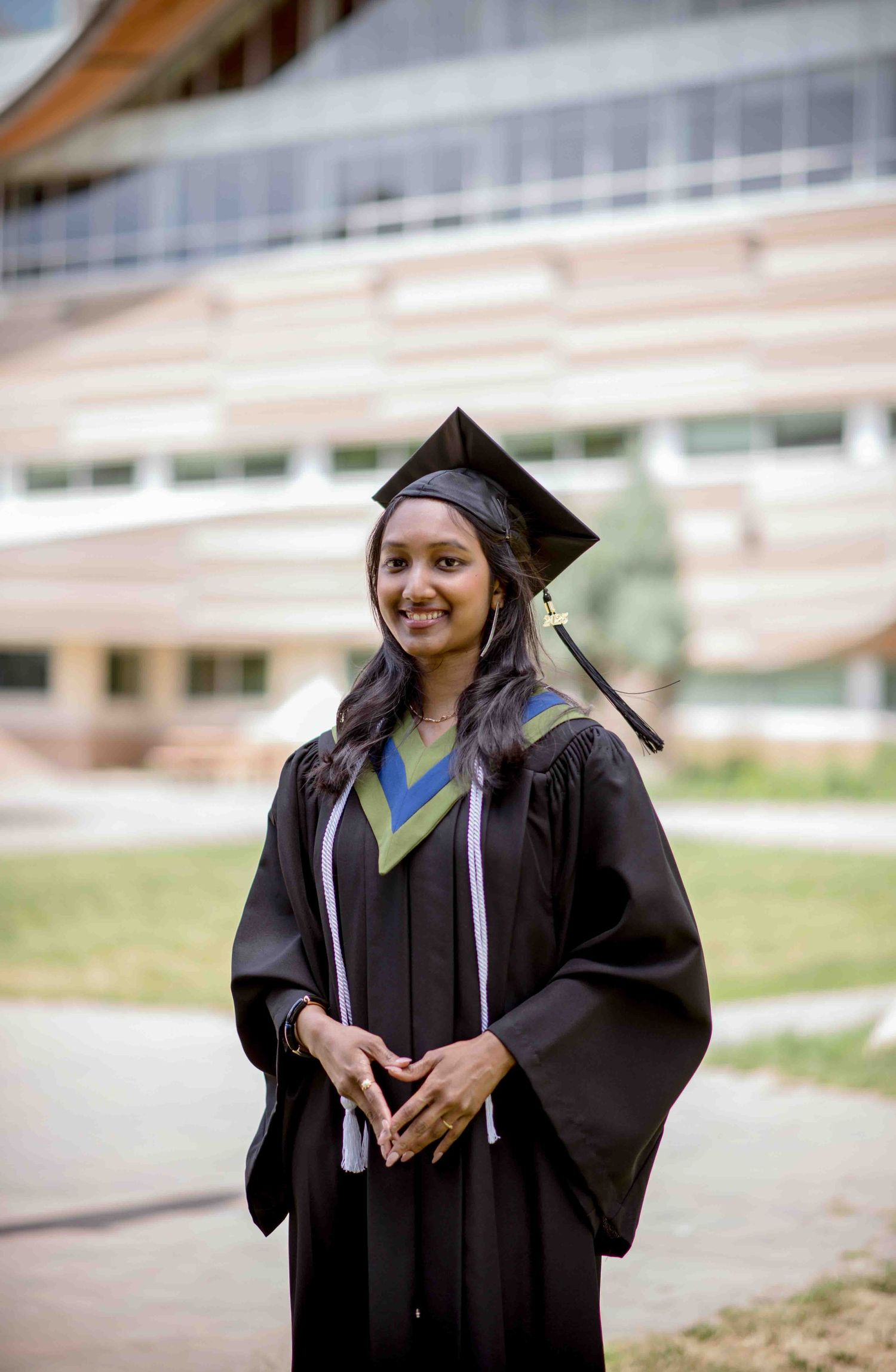 Smiling graduate in black cap and gown stands proudly outside modern university building.