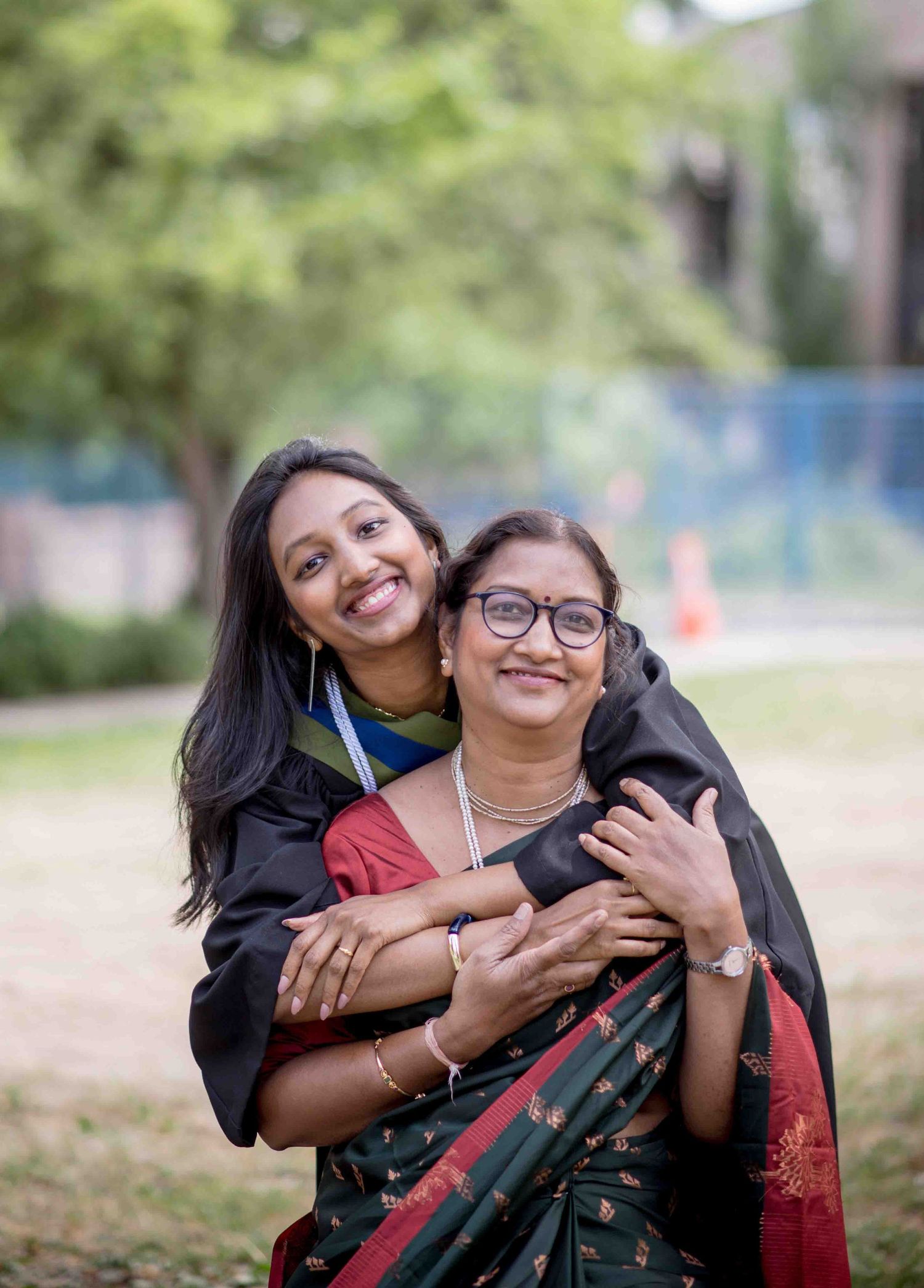 Graduate in black gown hugs smiling woman in traditional Indian saree outdoors at university campus.