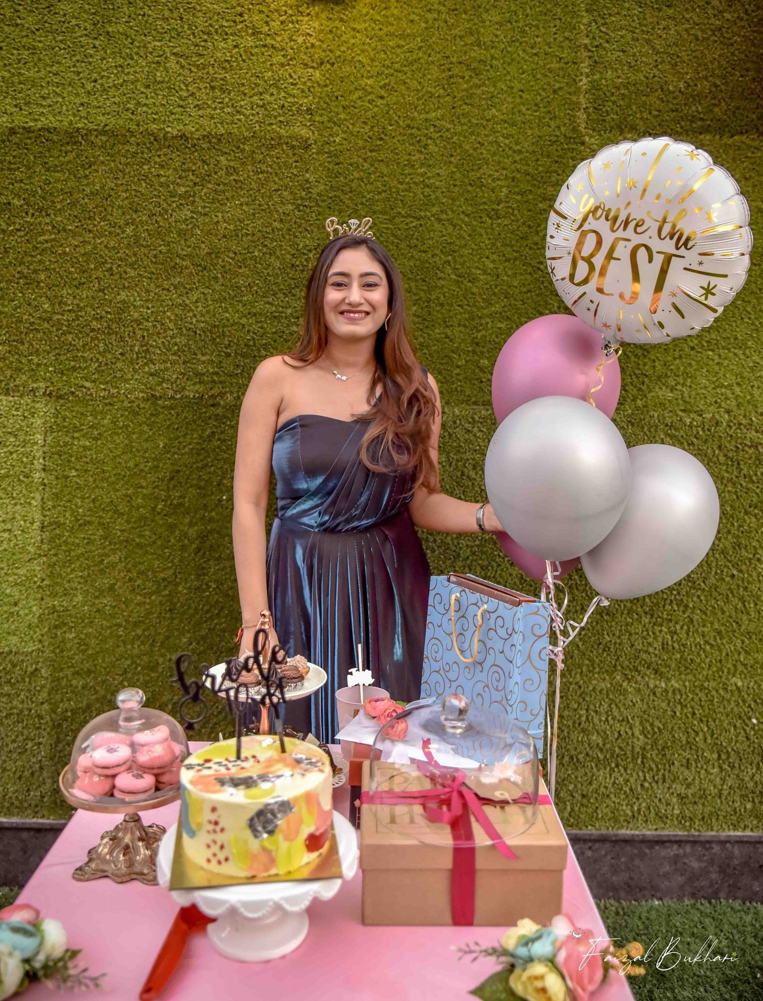 Bride-to-be in blue dress stands by decorated dessert table with cake, balloons and gifts at bridal shower.