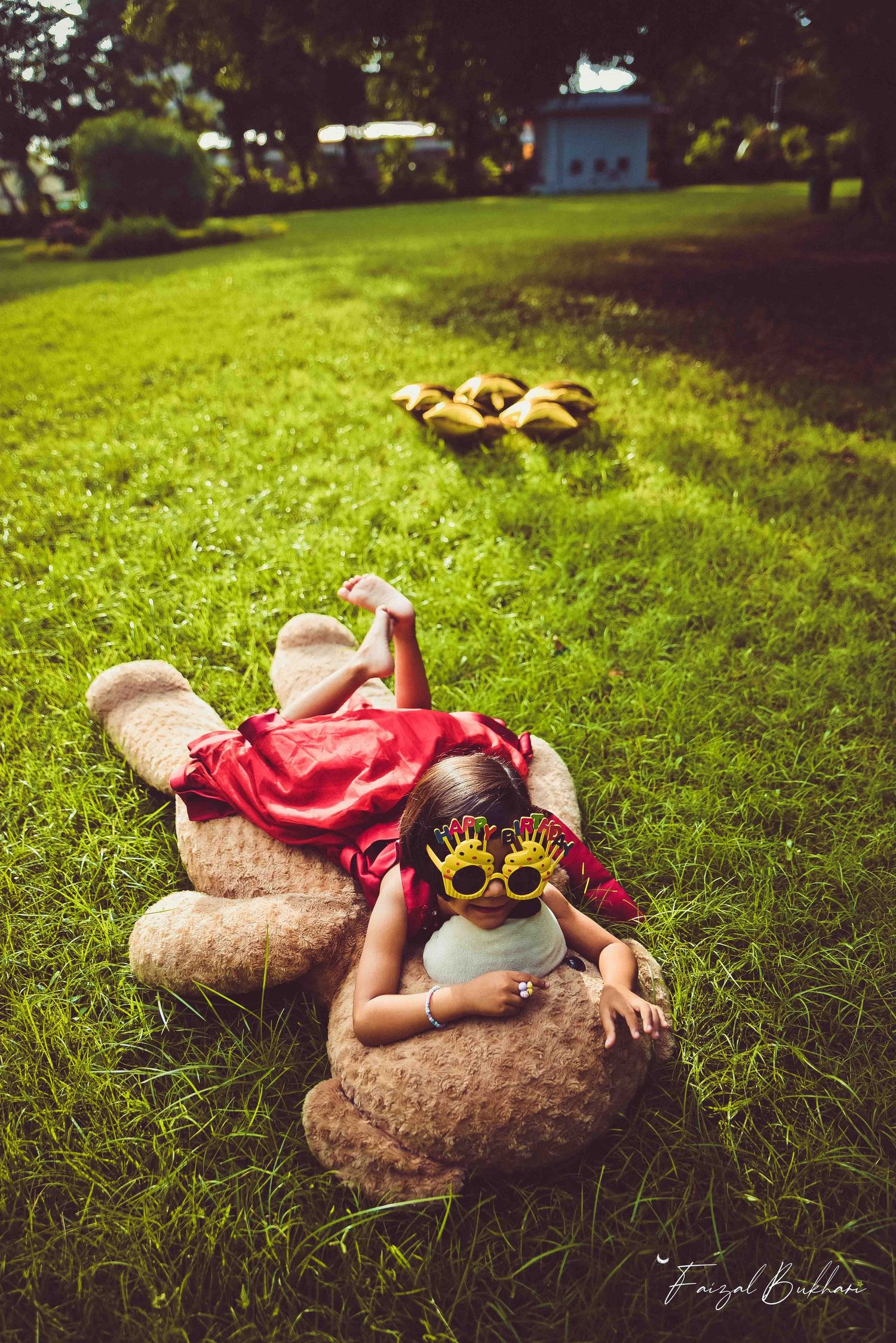 A young girl in red dress playing on a large teddy bear on green grass wearing novelty glasses.