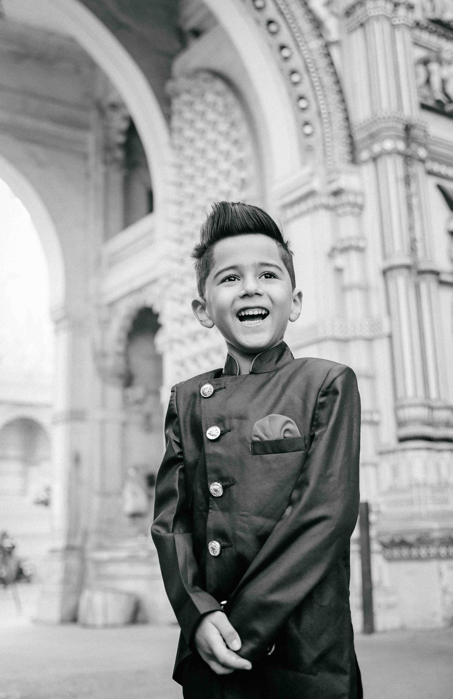 Laughing boy in black Nehru suit with stylish mohawk haircut standing before ornate architectural arches.