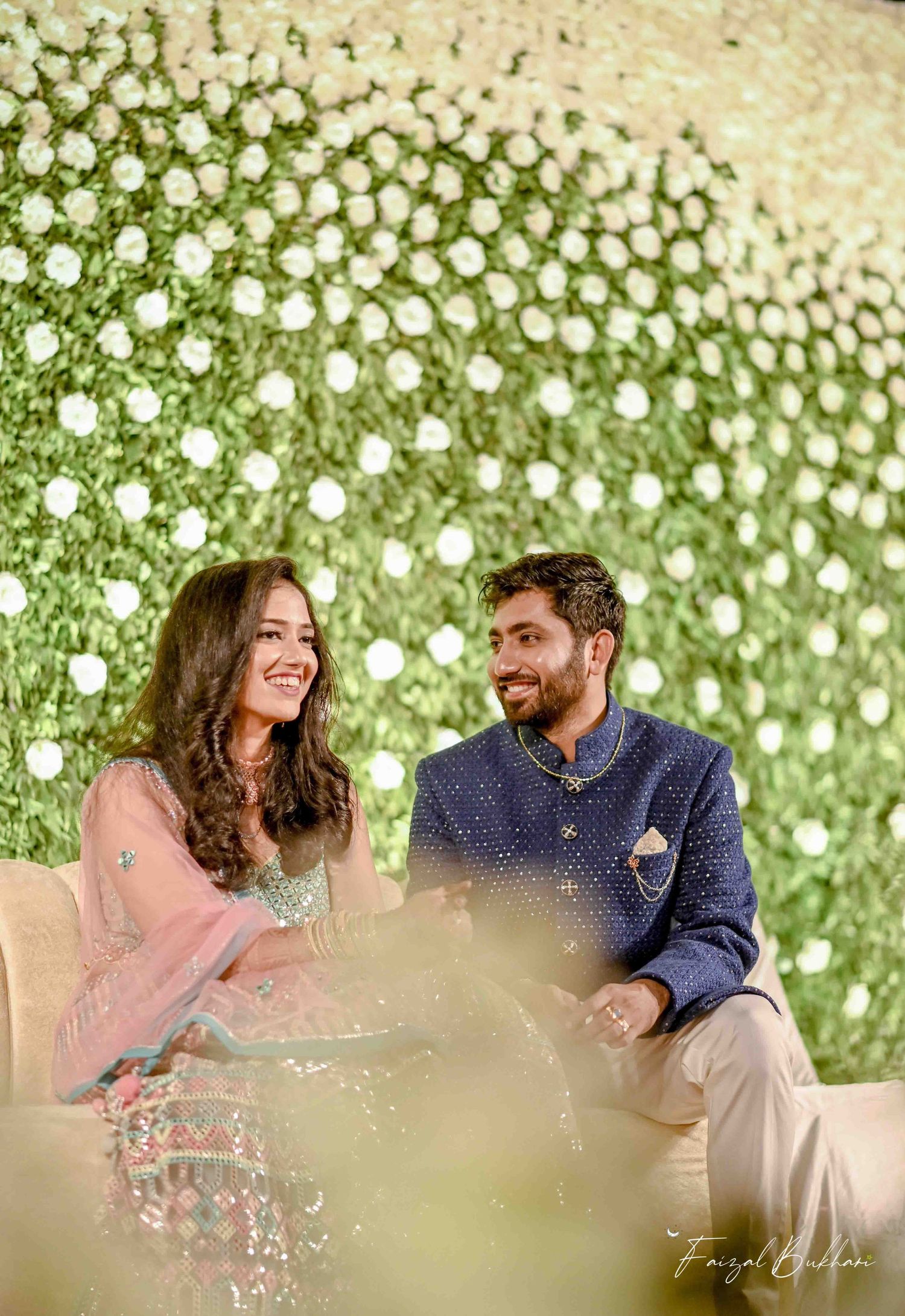 A happy Indian couple sits together laughing in front of a stunning white floral wall backdrop at their engagement.