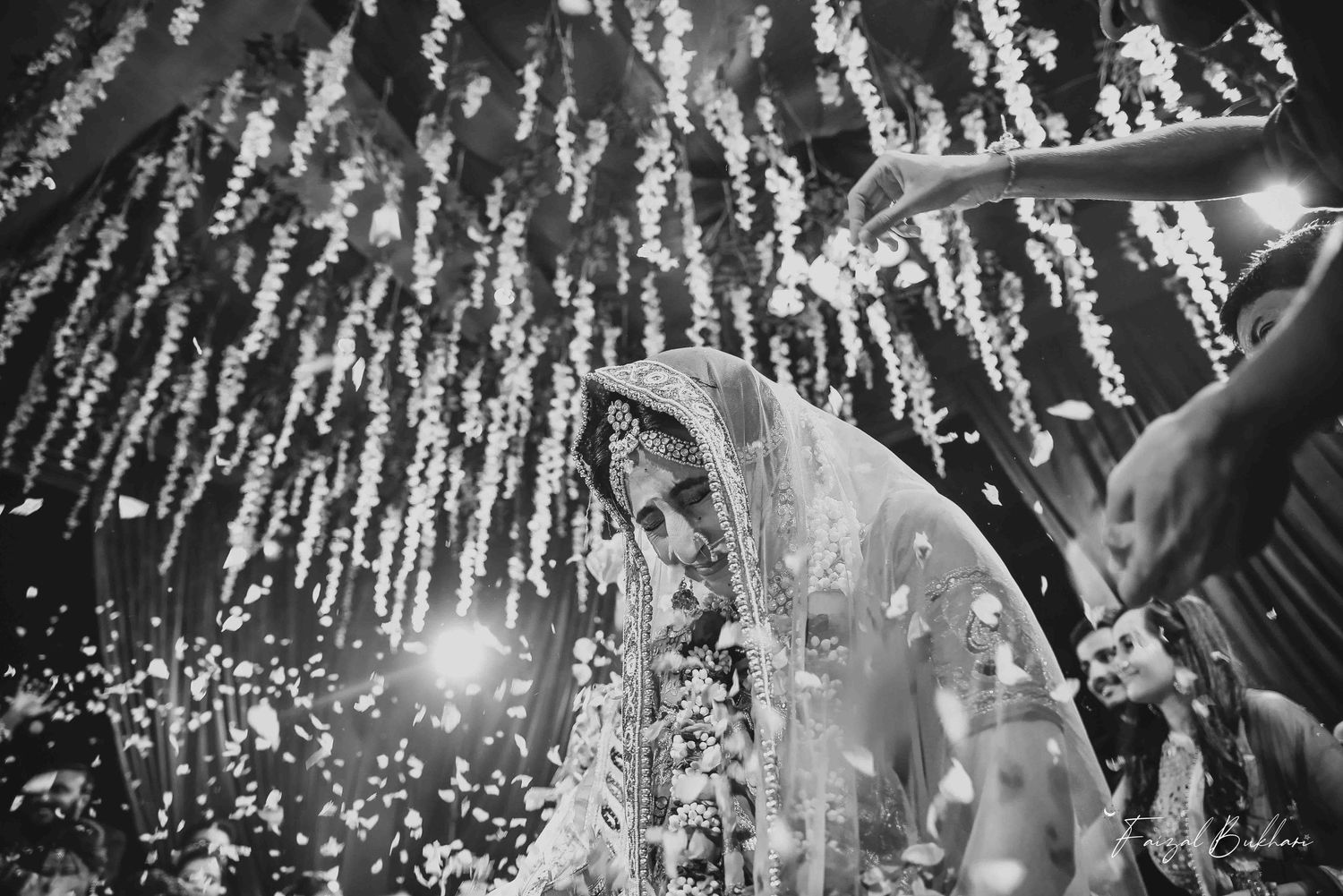 Black and white photo of Indian bride in ornate veil surrounded by falling flowers at wedding ceremony.