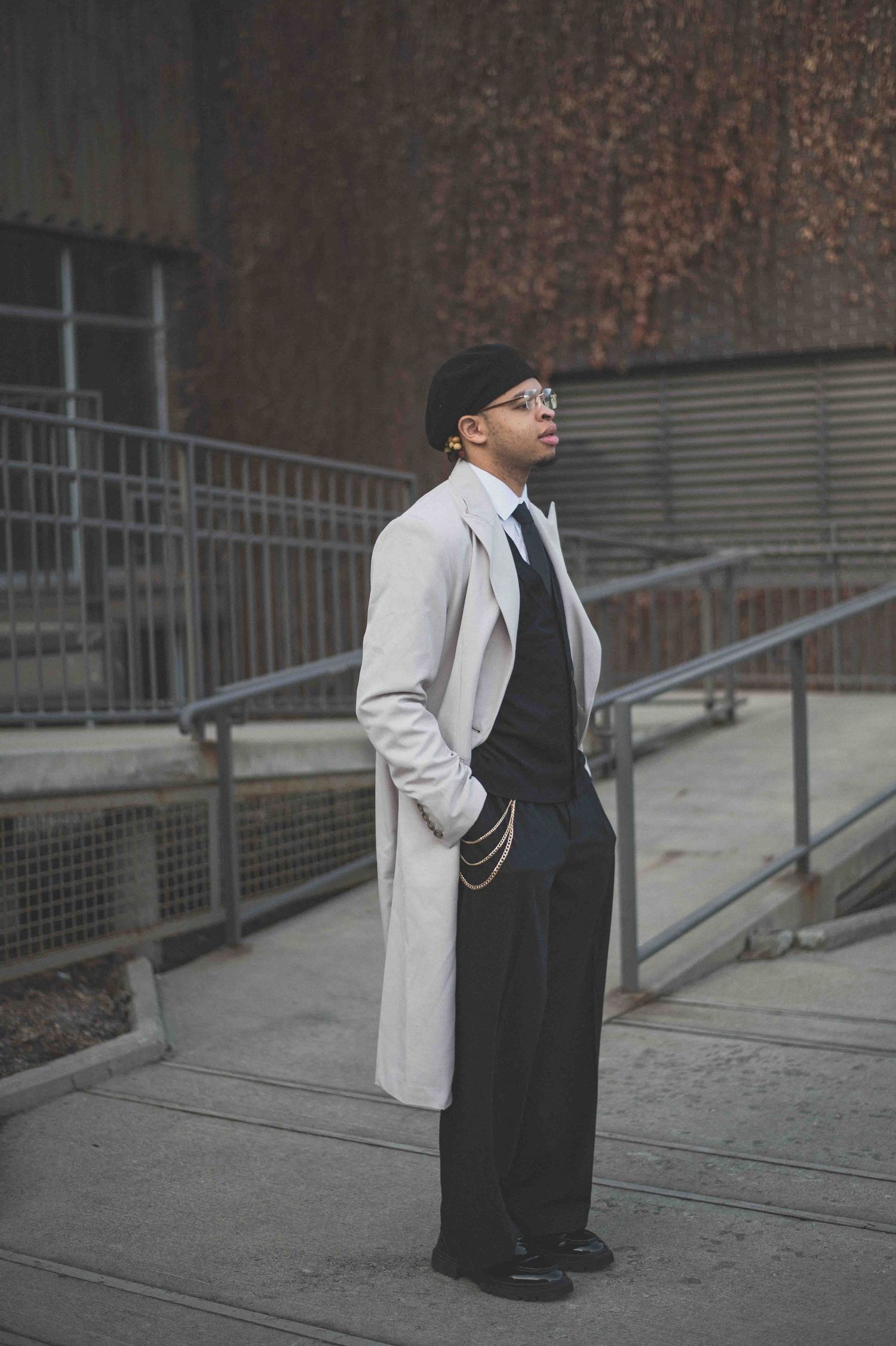 Stylish man in white trench coat, black hat, and wide-leg trousers posing outdoors against rustic wall.