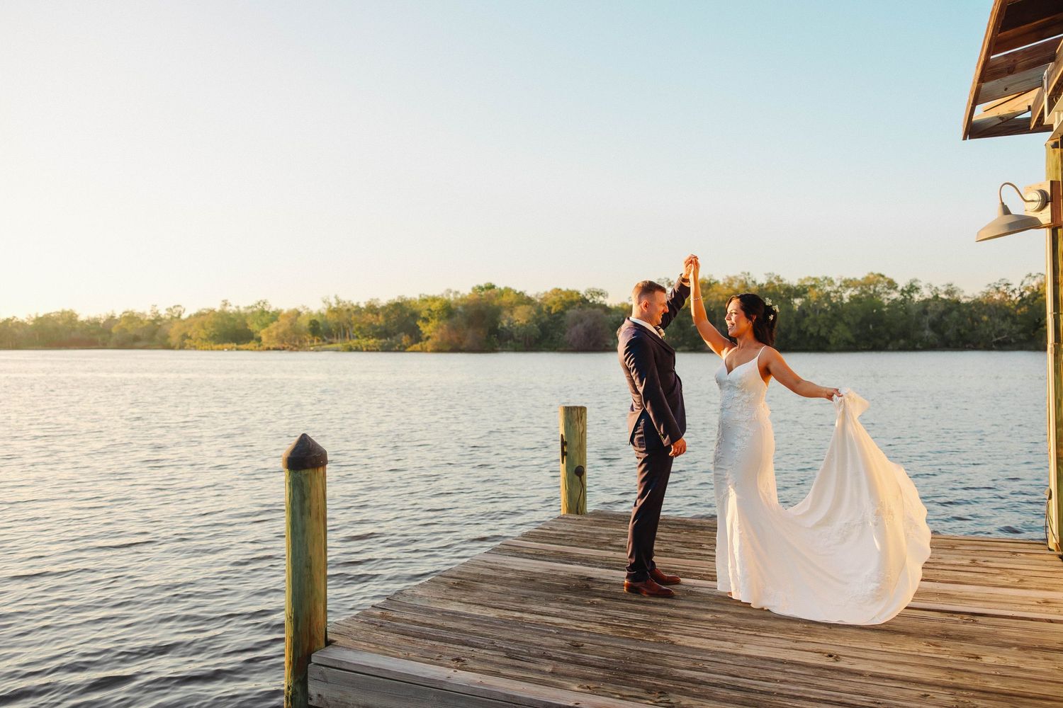 Bride and groom dancing on a wooden dock at sunset by a serene lake, bride's white gown flowing in the breeze.