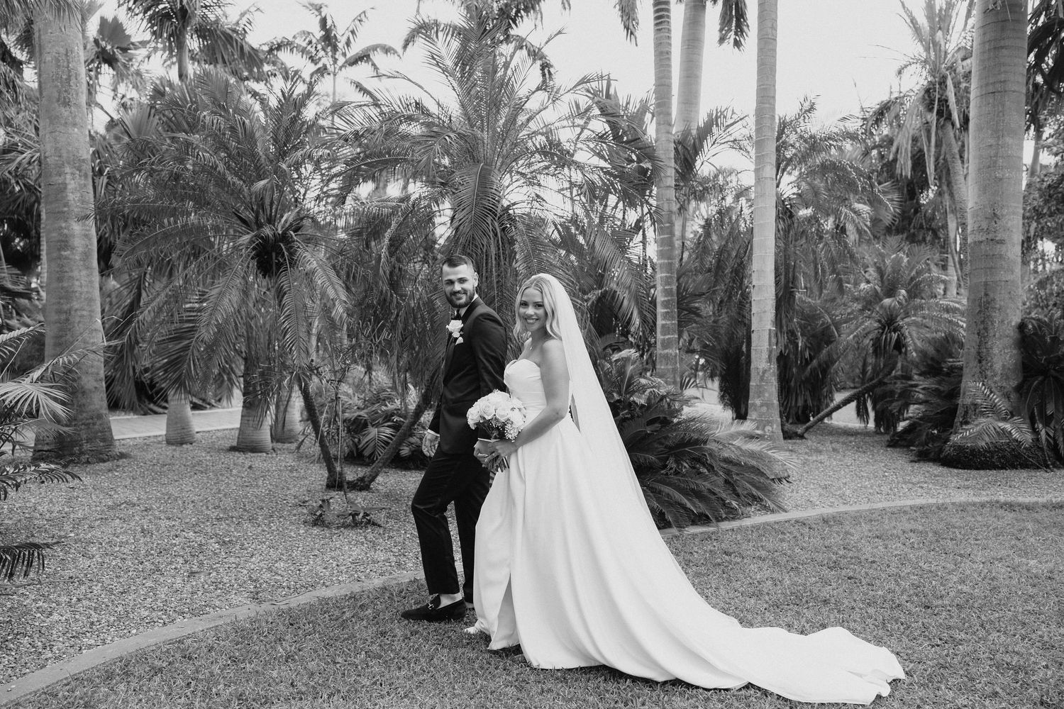 A bride and groom walk hand in hand among tropical palm trees in a classic black and white wedding photo.