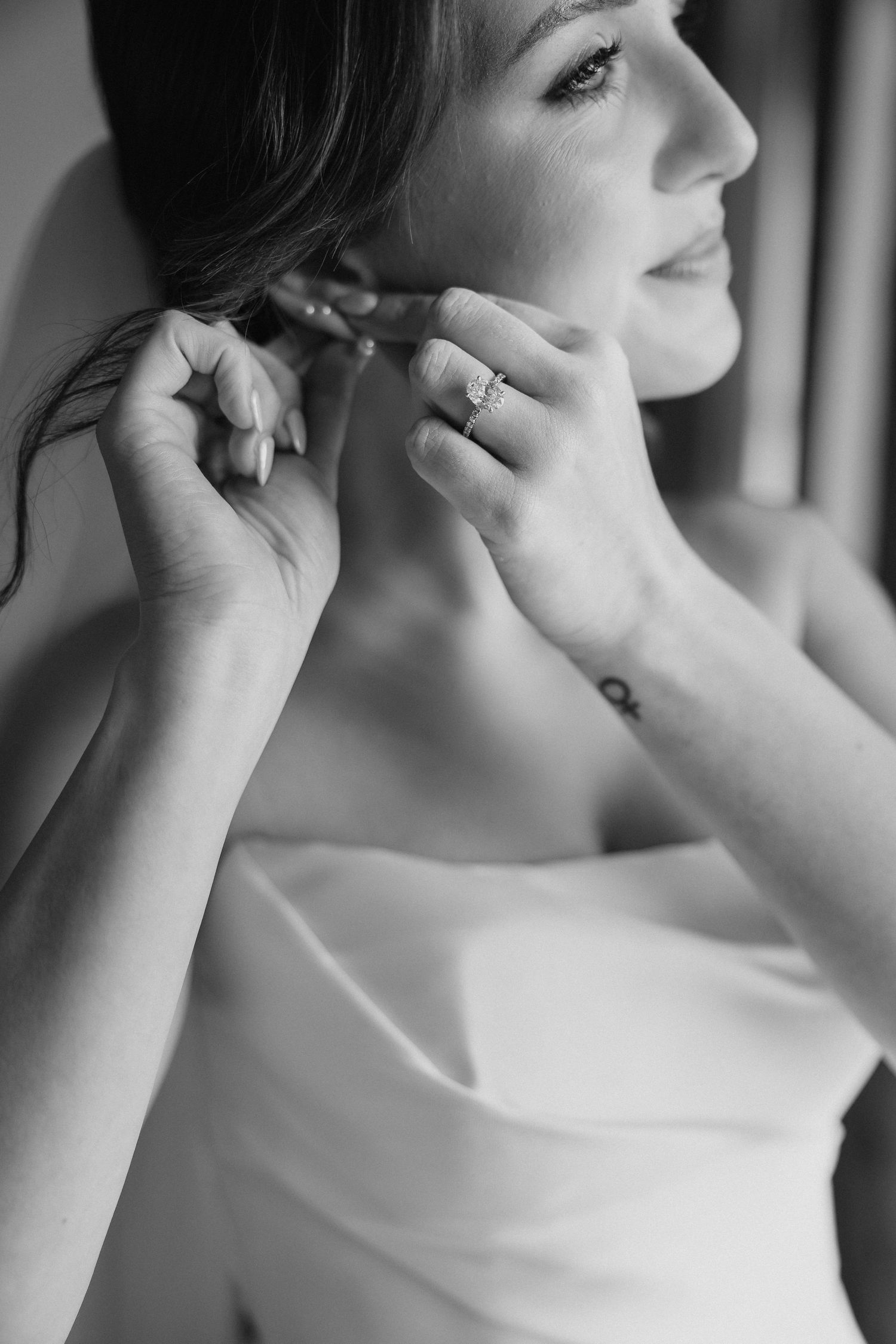 Black and white photo of a bride putting on earrings, wearing a strapless gown and diamond engagement ring.