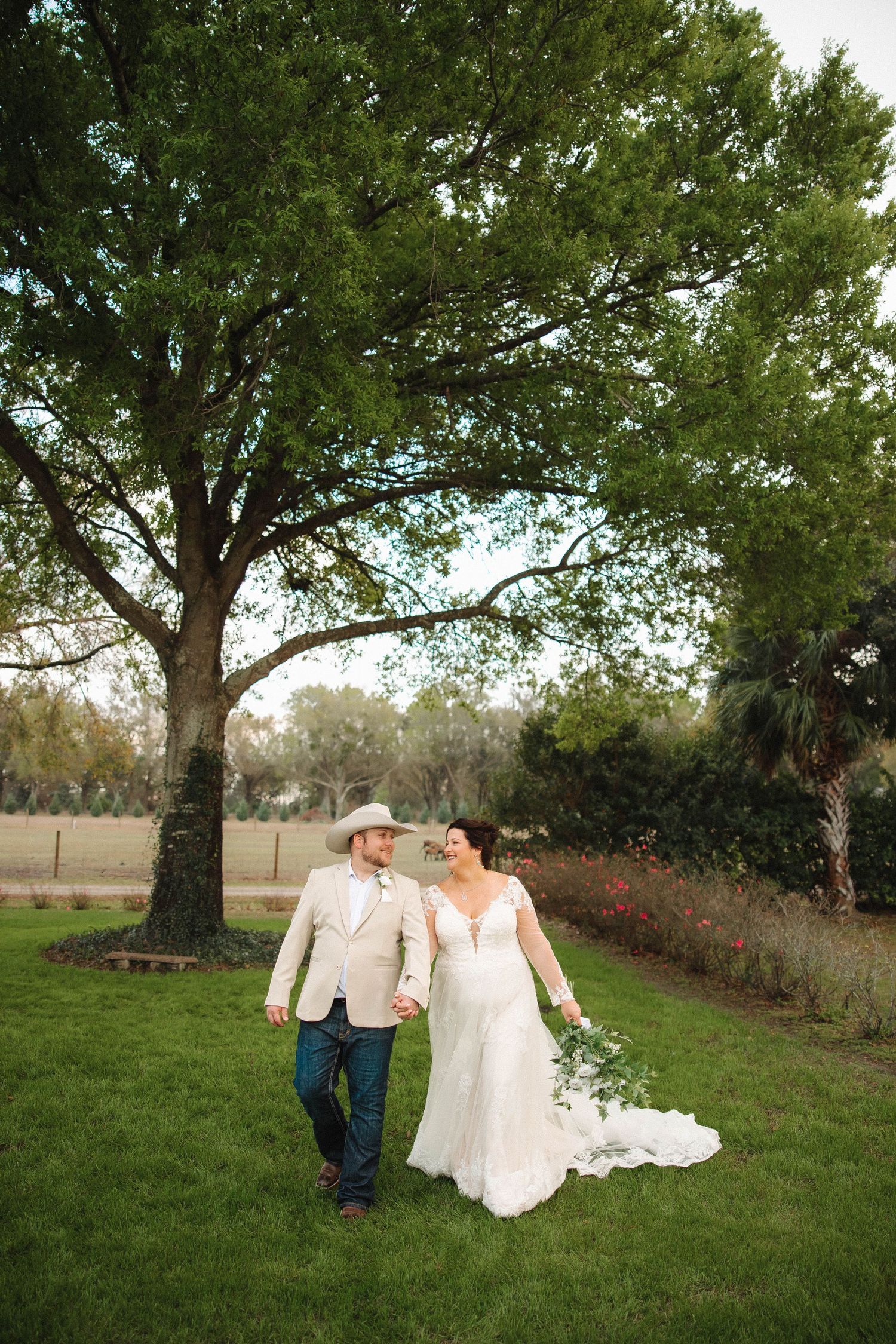 Bride and groom walk hand-in-hand on lush green lawn beneath a large oak tree at an outdoor wedding venue.