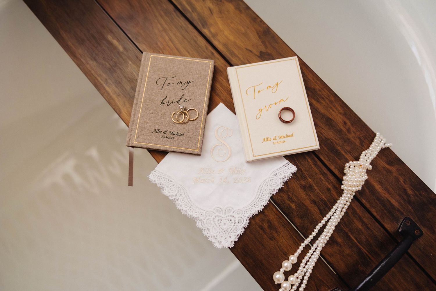 Wedding vow books, gold rings, lace handkerchief, and pearl necklace displayed on a wooden tray.
