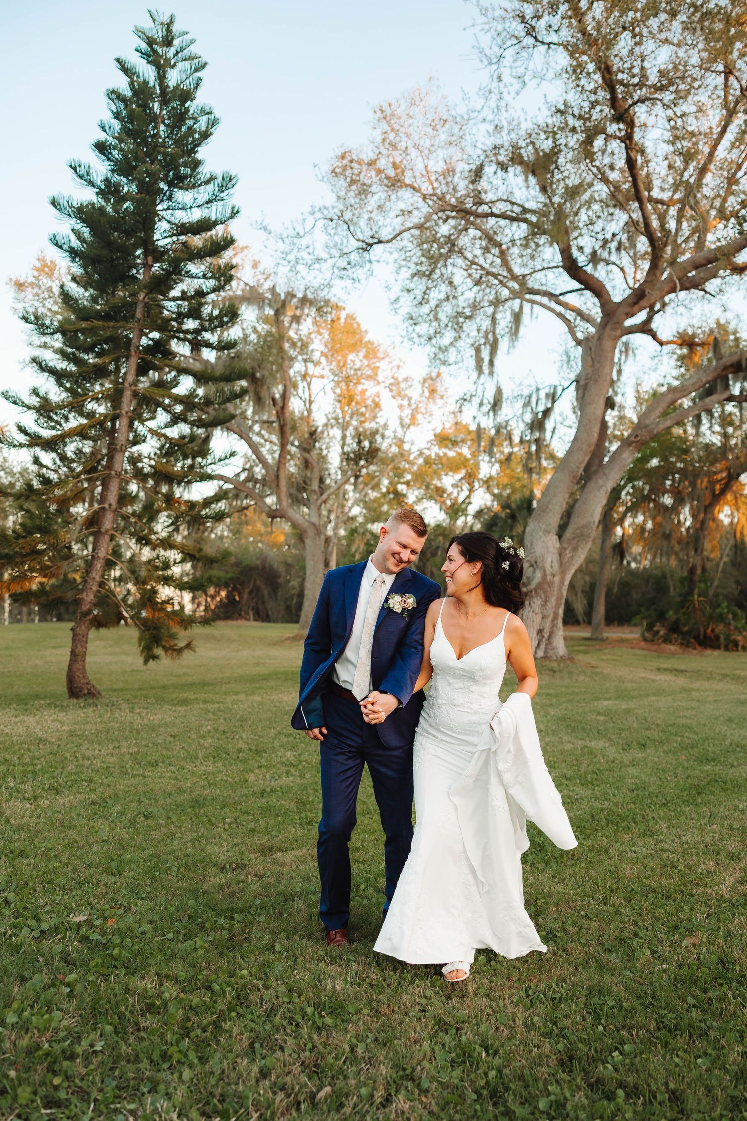 Bride and groom walk hand-in-hand through lush green park at golden hour, she in white gown, he in navy suit.