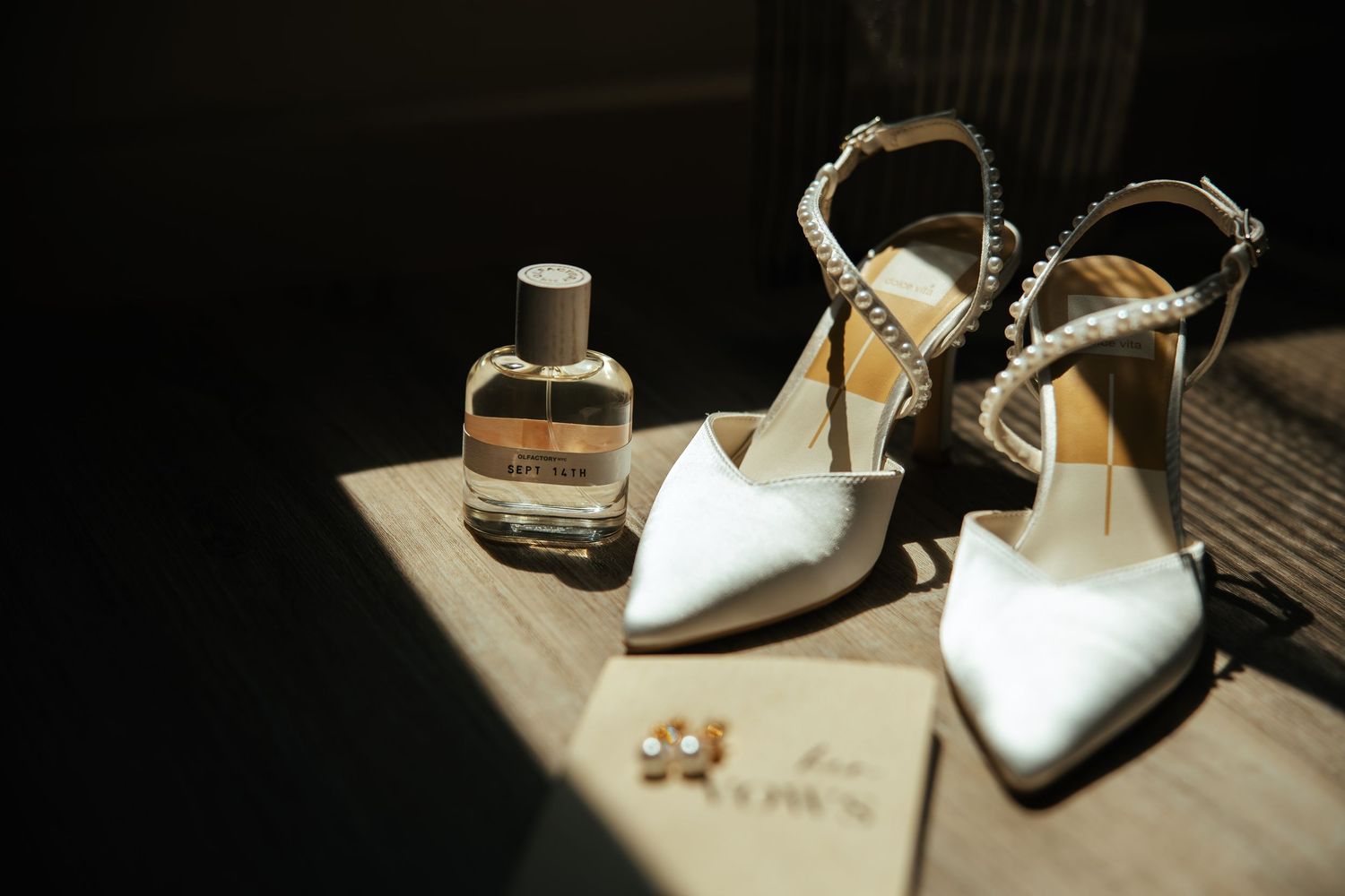 White bridal heels with crystal straps beside a perfume bottle and earrings on a wooden surface in soft light.