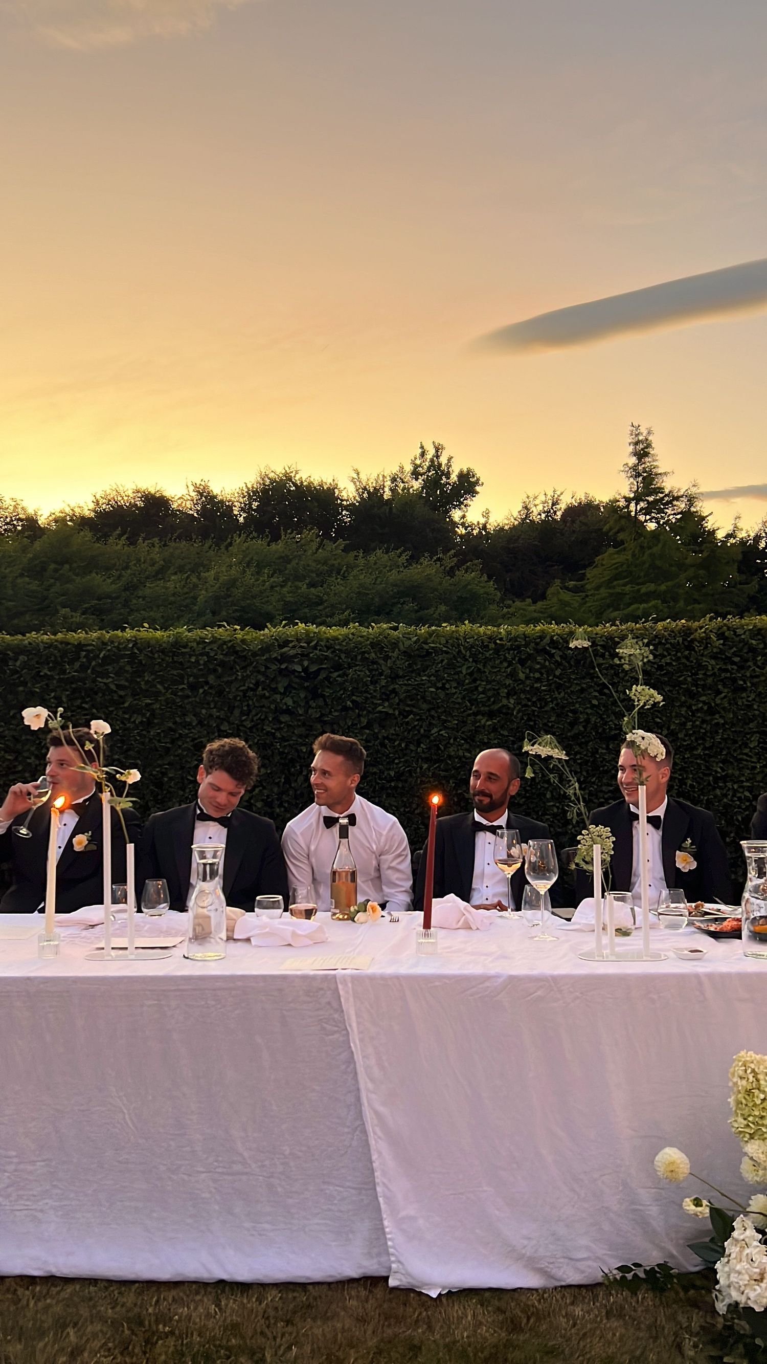 Long banquet table with white tablecloth set against sunset sky with guests seated for outdoor wedding dinner.