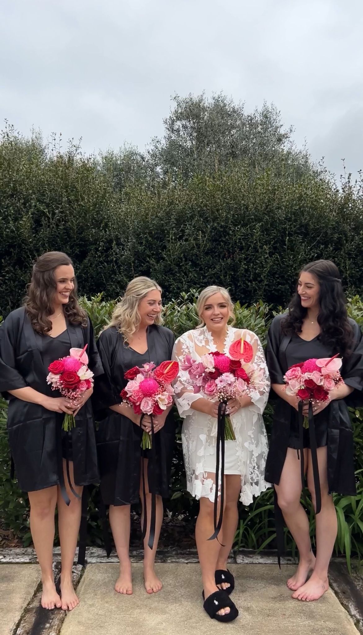 Bridesmaids in black dresses holding vibrant pink bouquets pose barefoot against garden hedge.