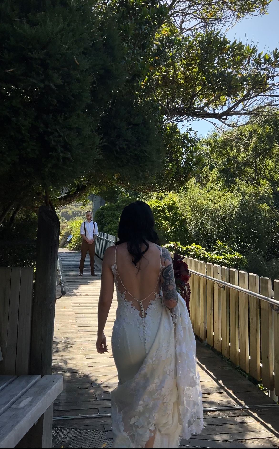 A couple walks along a wooden boardwalk path surrounded by lush green trees and vegetation on a sunny day.