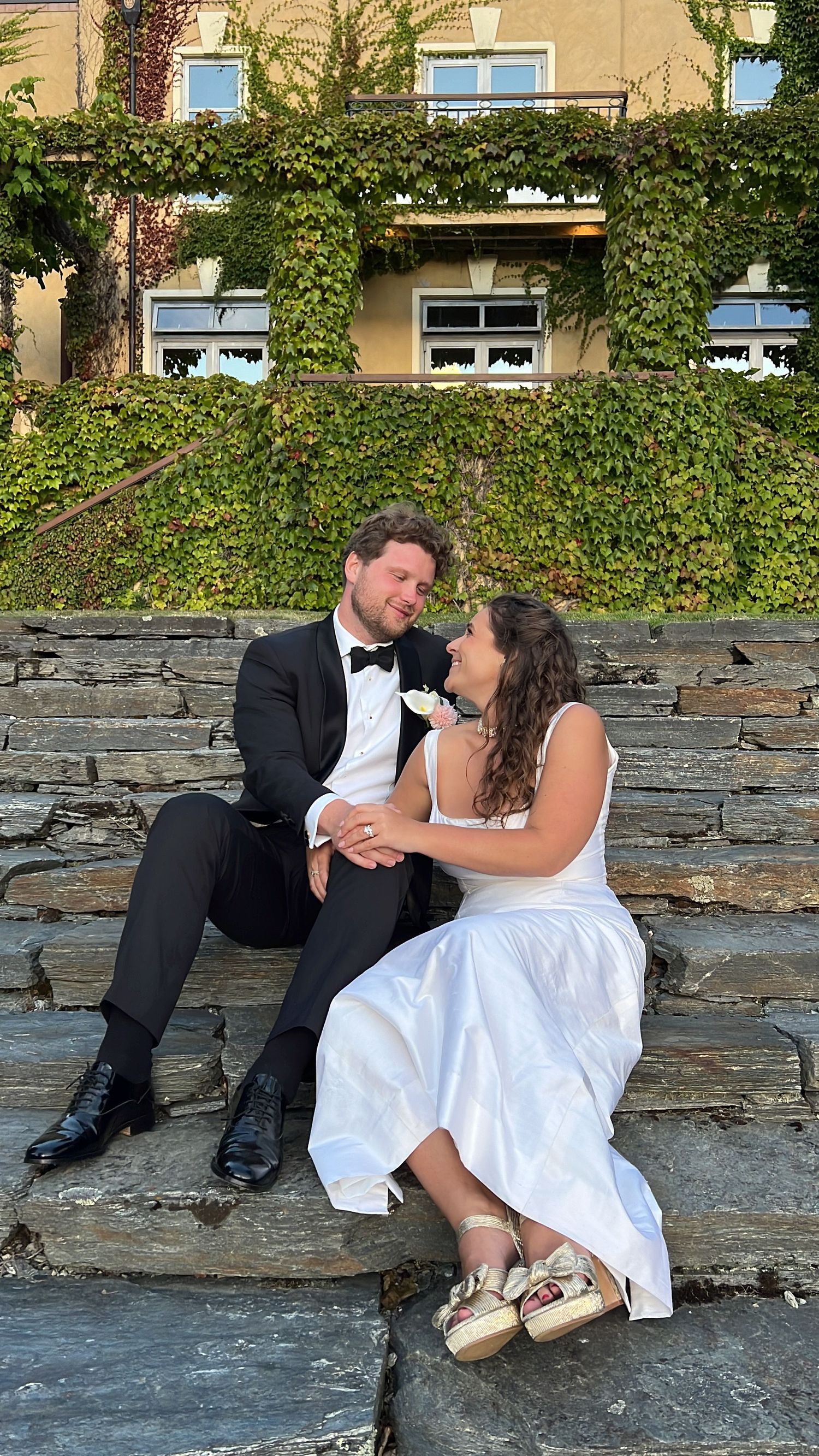 Wedding couple sitting on stone wall with ivy-covered building in background.