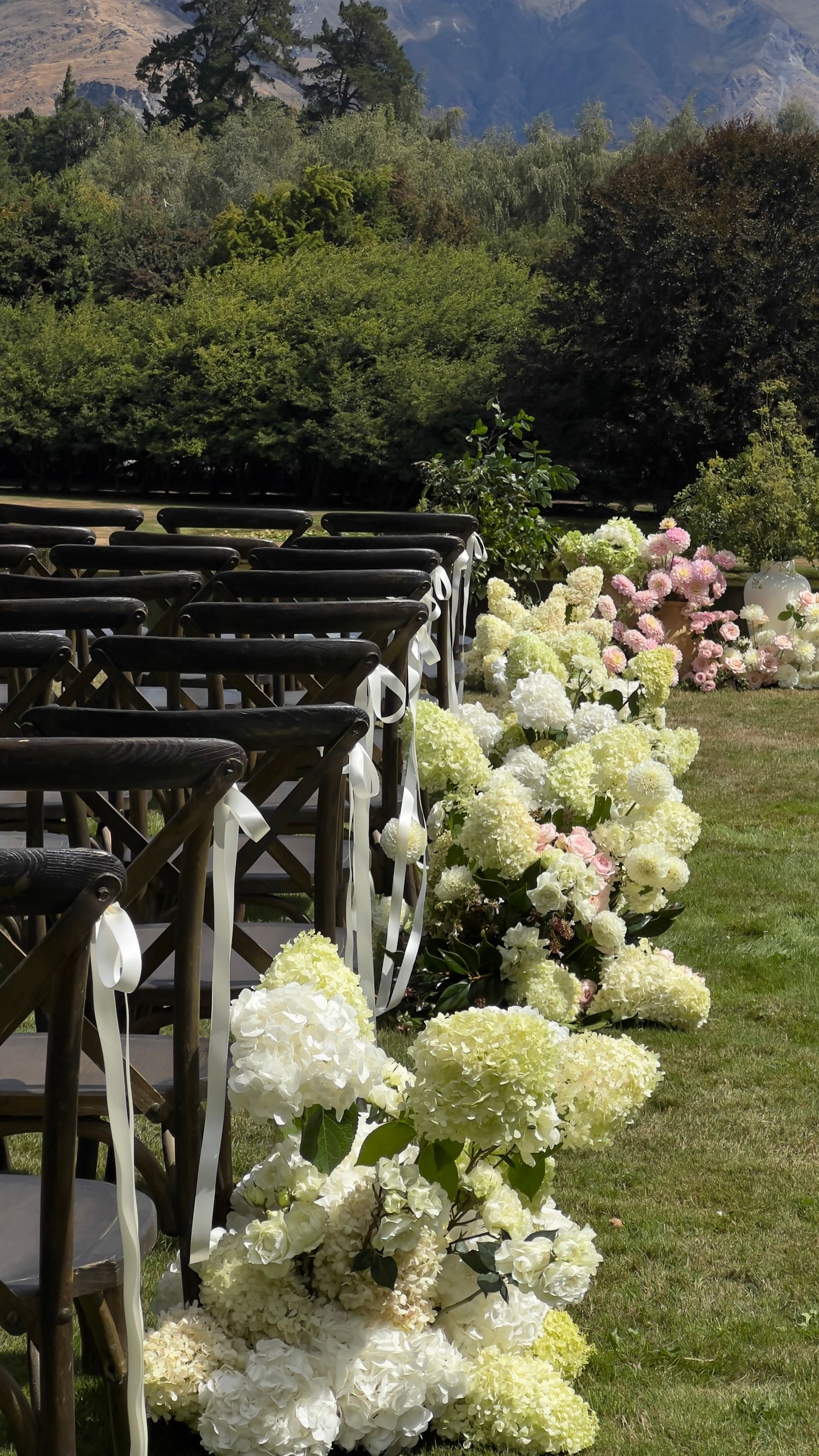 Wedding ceremony setup with white hydrangea arrangements decorating wooden chairs against mountain scenery. content creation in new zealand