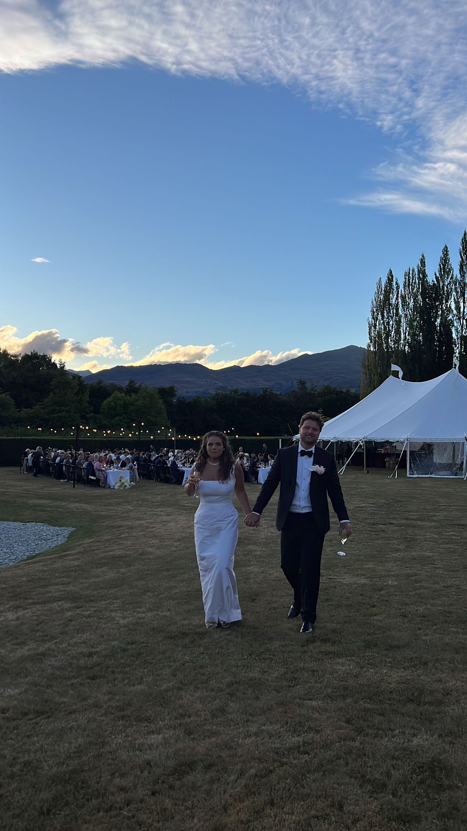 Wedding couple walking across a grassy field at dusk with mountains and event tent in background.