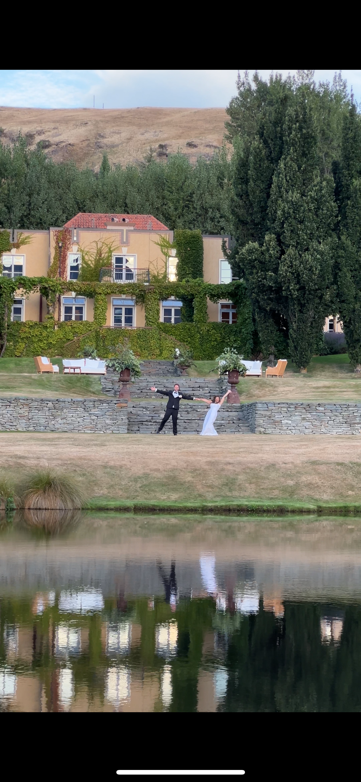 A wedding photo session taking place by a serene lake with buildings and trees in the background, creating reflections on water.