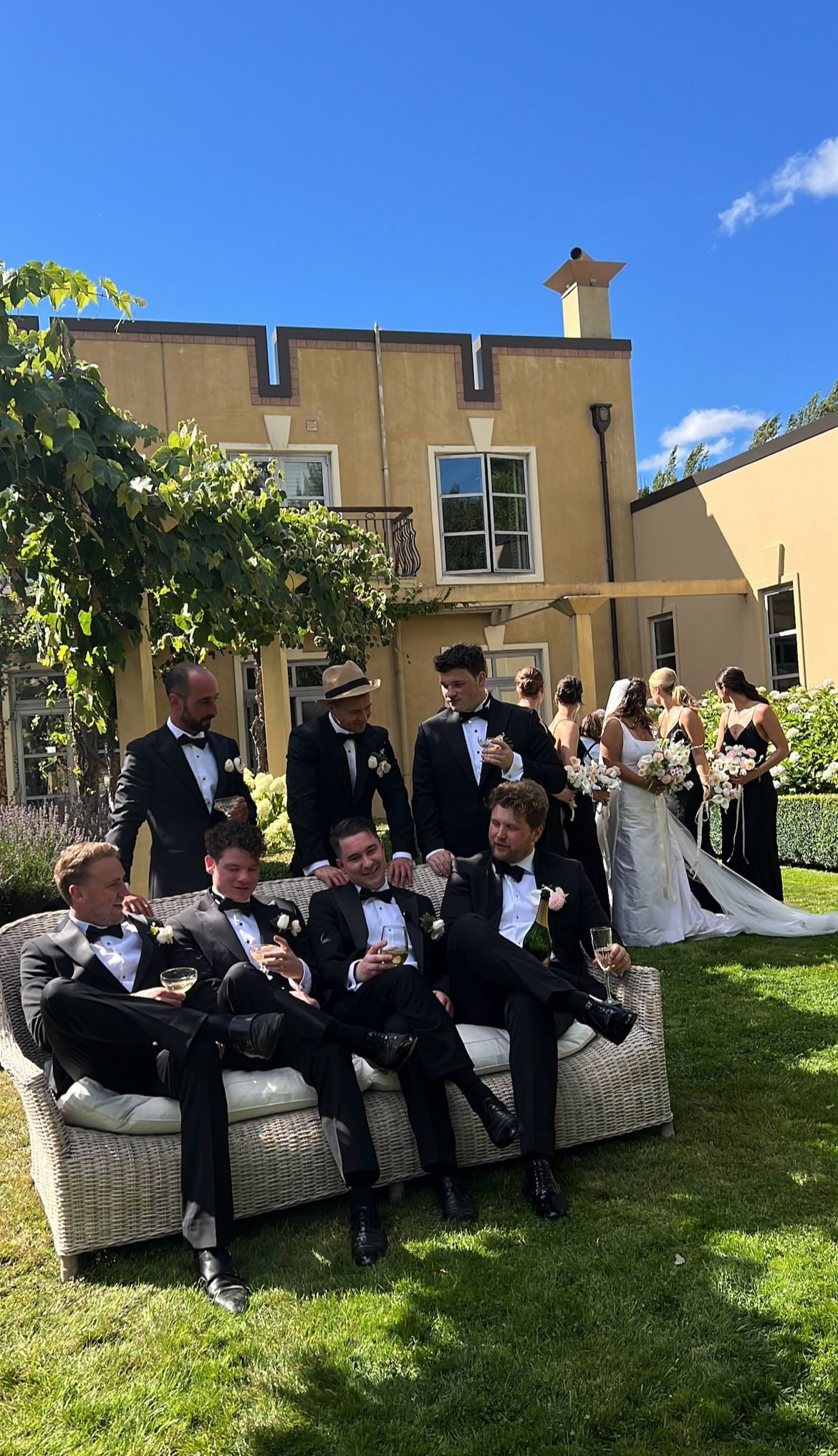 Groomsmen in black tuxedos relax on white wicker outdoor furniture during a wedding celebration on a sunny day.
