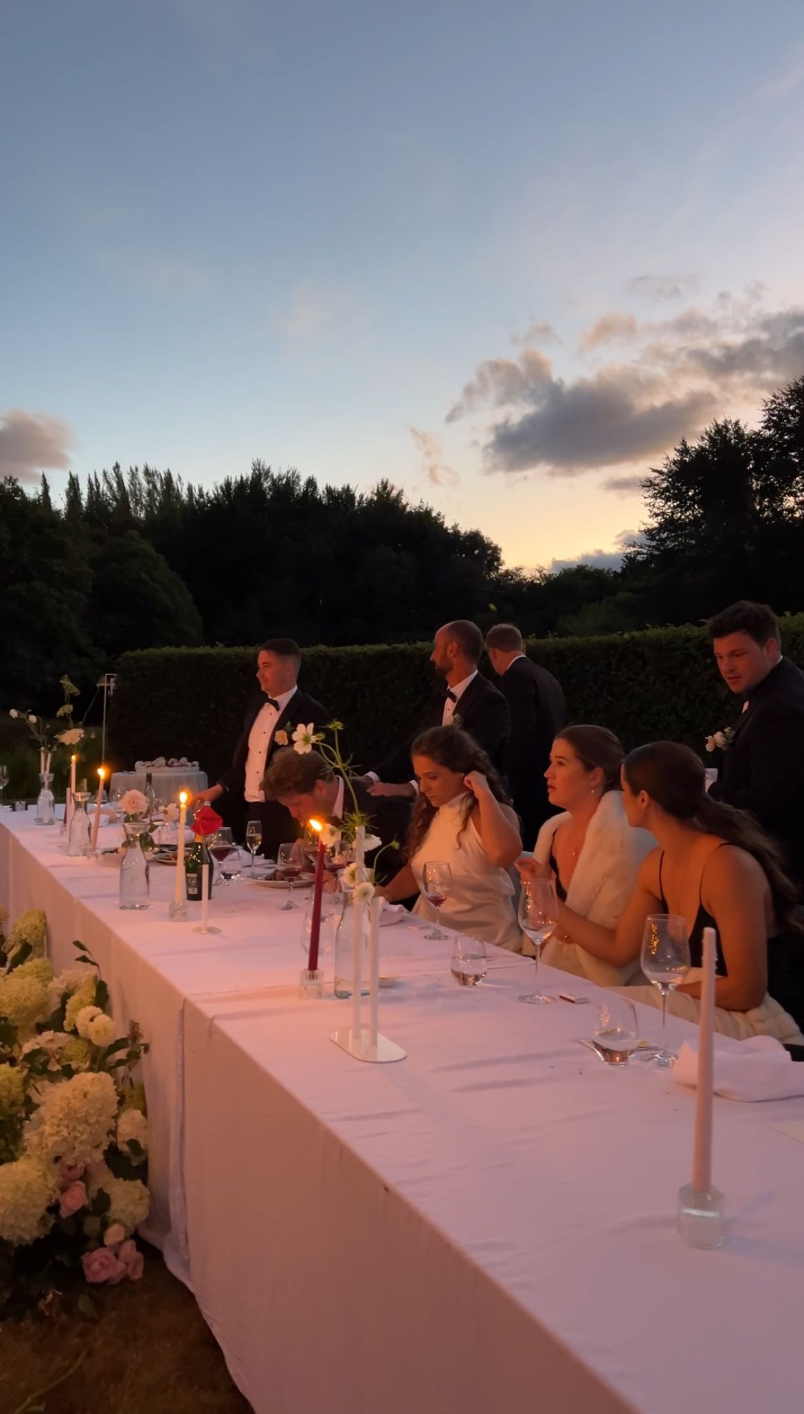 Wedding party guests seated at an elegant white dining table during sunset at an outdoor reception celebration.
