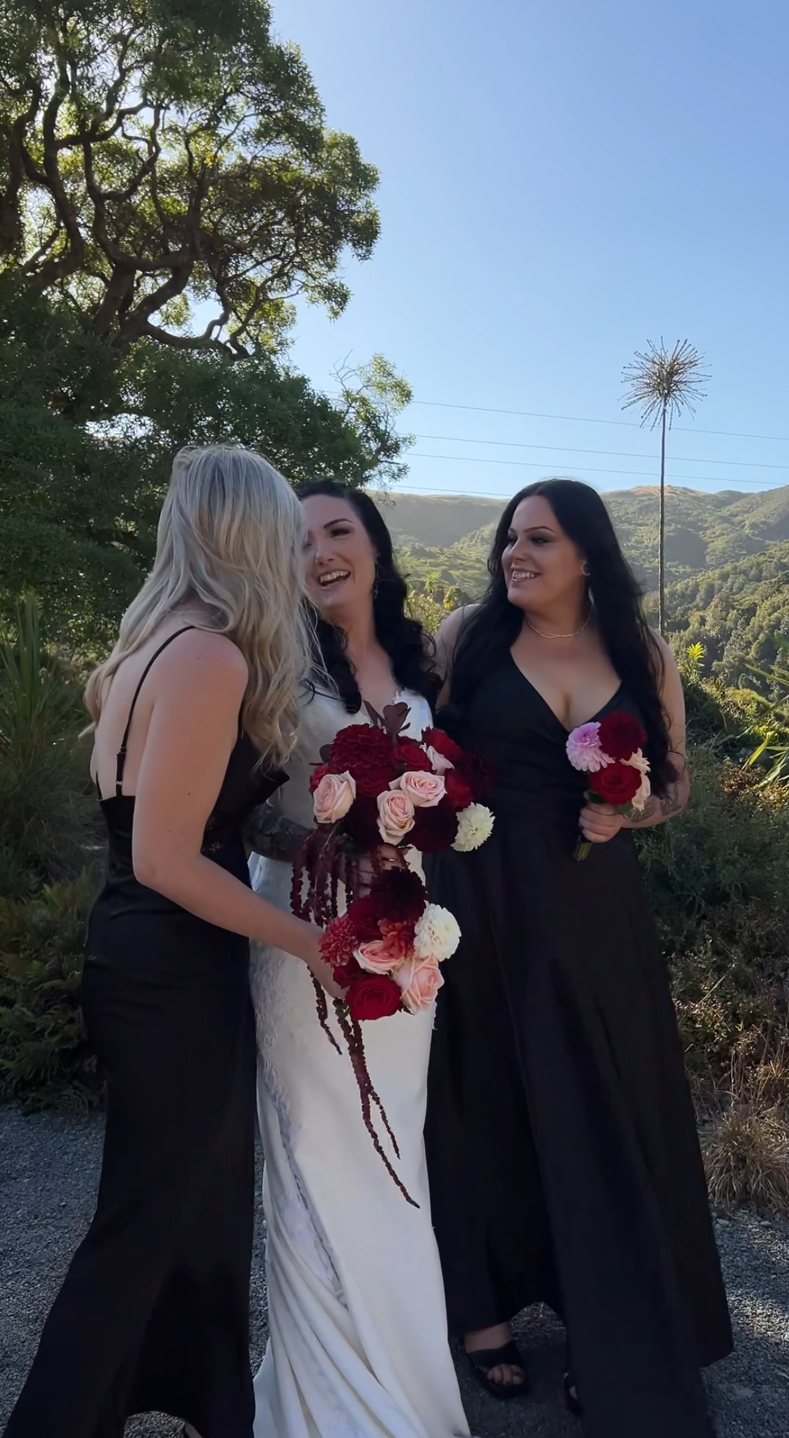 Three people in formal black and white attire share laughter while holding red and white floral bouquets outdoors.
