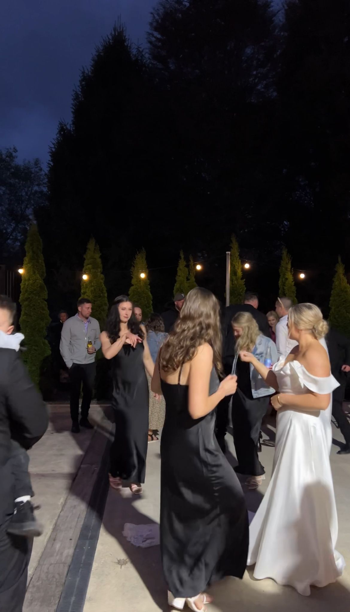 Wedding guests dance together on an outdoor patio at night with illuminated lamp posts in the background.