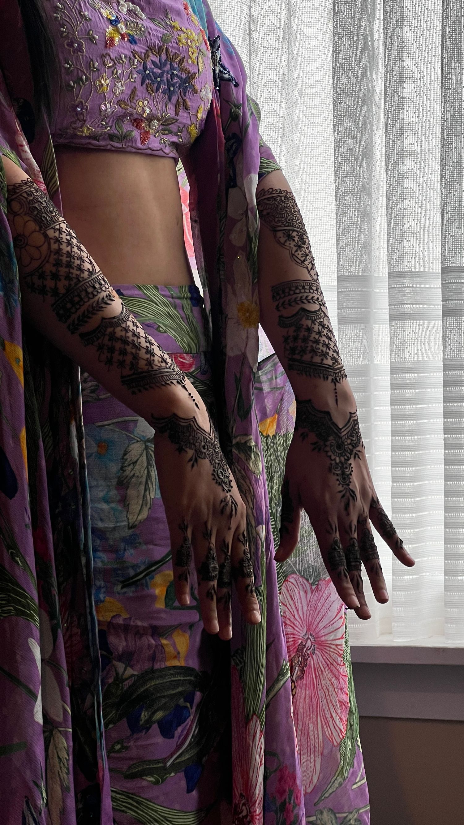 Intricate henna designs adorn hands against a floral purple dress with pink flowers near vertical window blinds.