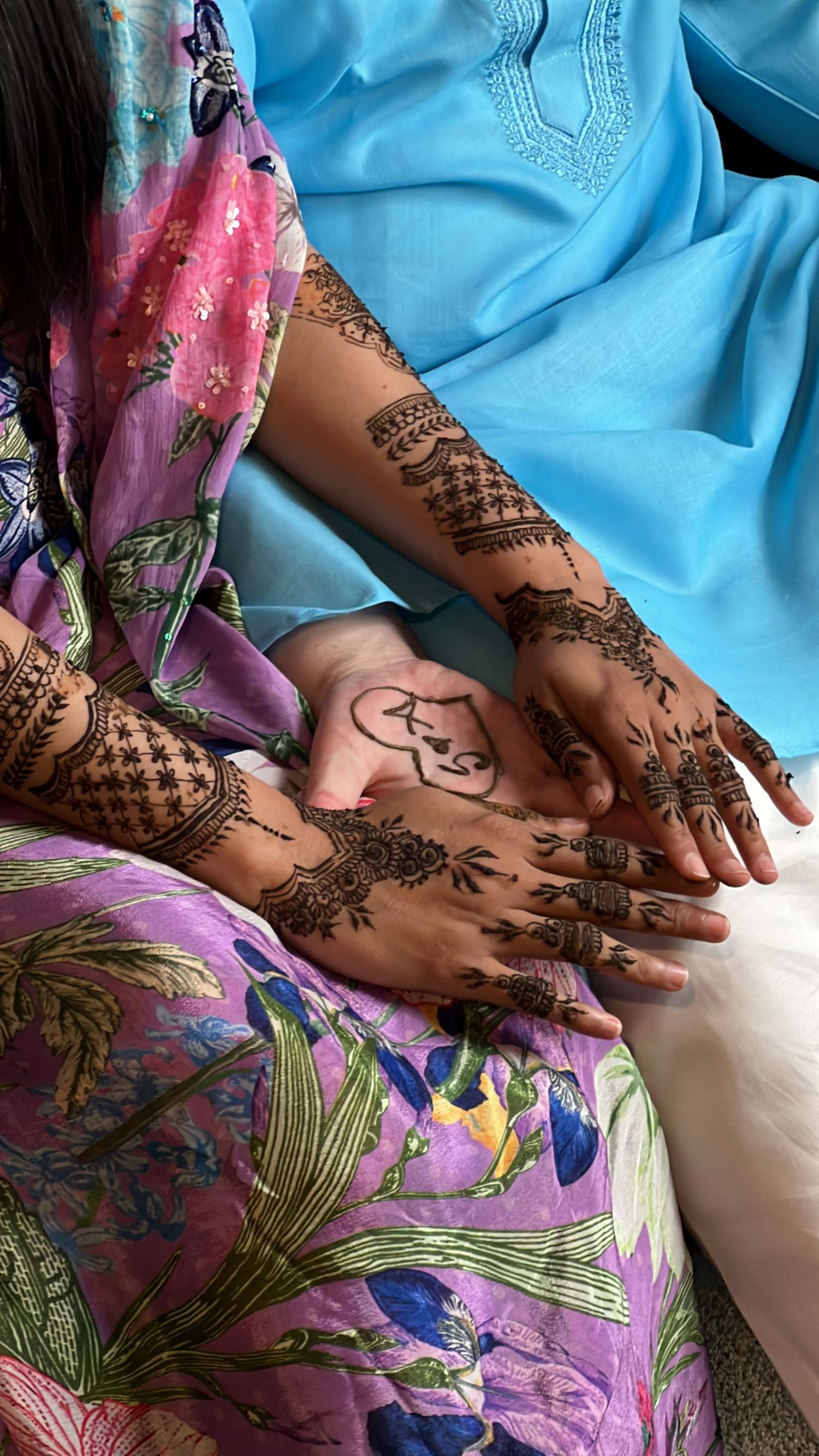 Ornate henna patterns decorating hands and arms against floral print fabric and blue clothing. henna night content new zealand