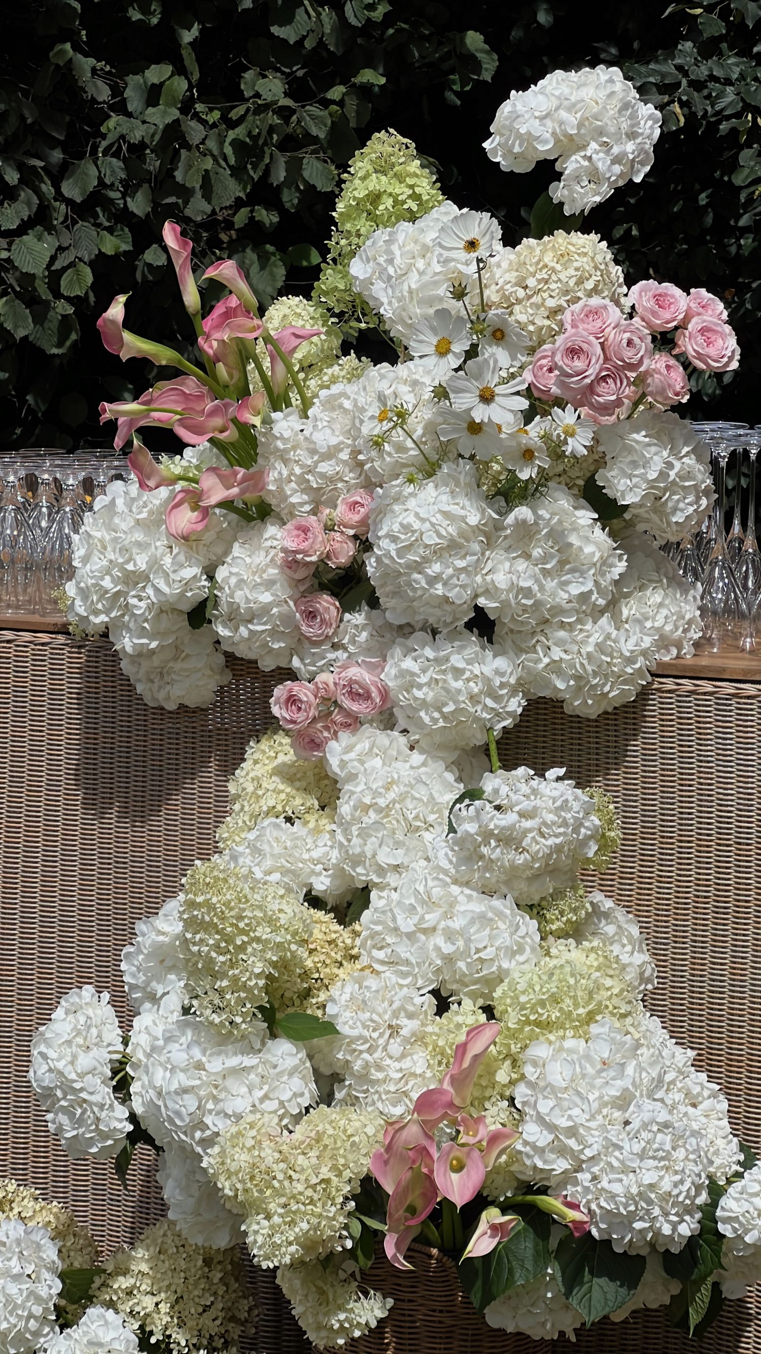 White hydrangeas and pink lilies arranged in an elegant floral display against wicker furniture.