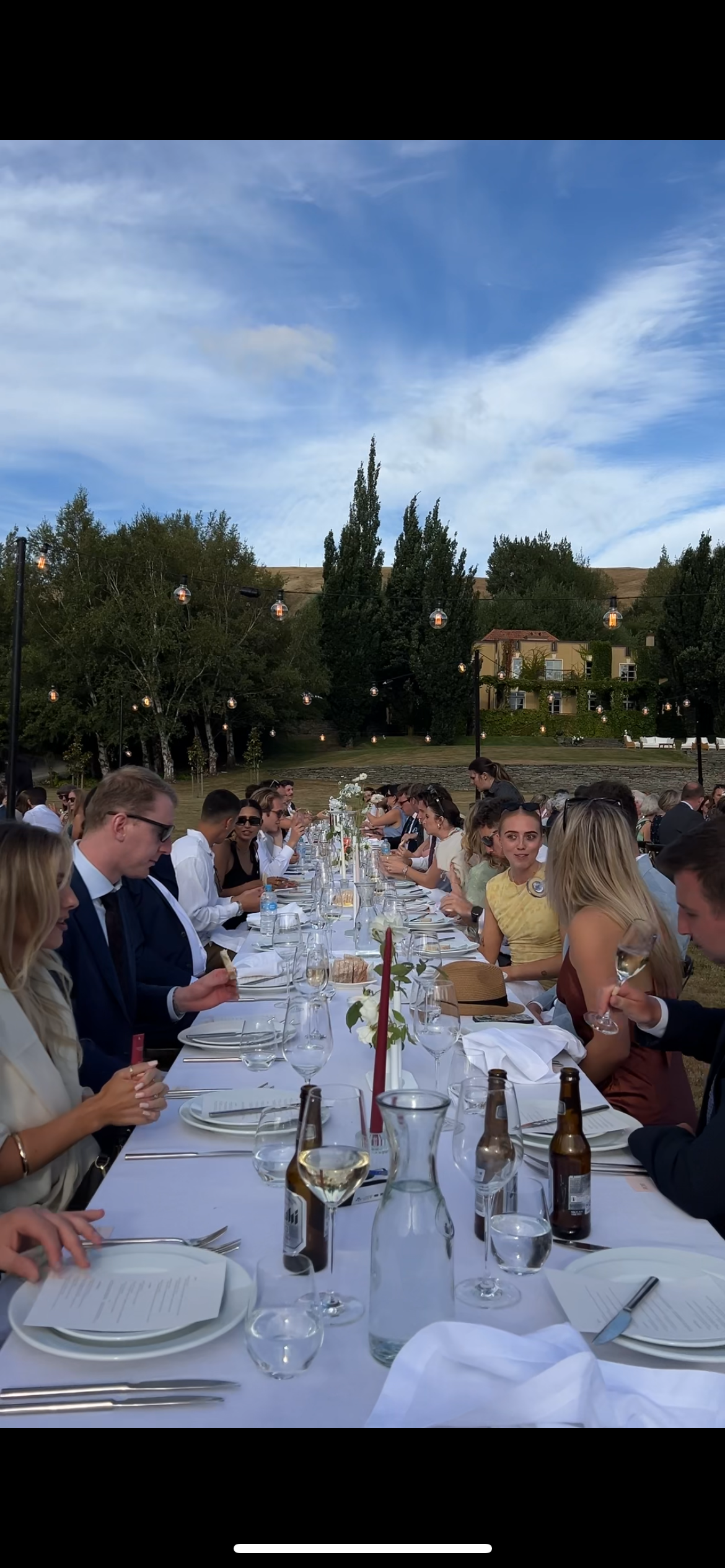 Long outdoor dining table set for a special event with guests seated under trees at sunset.