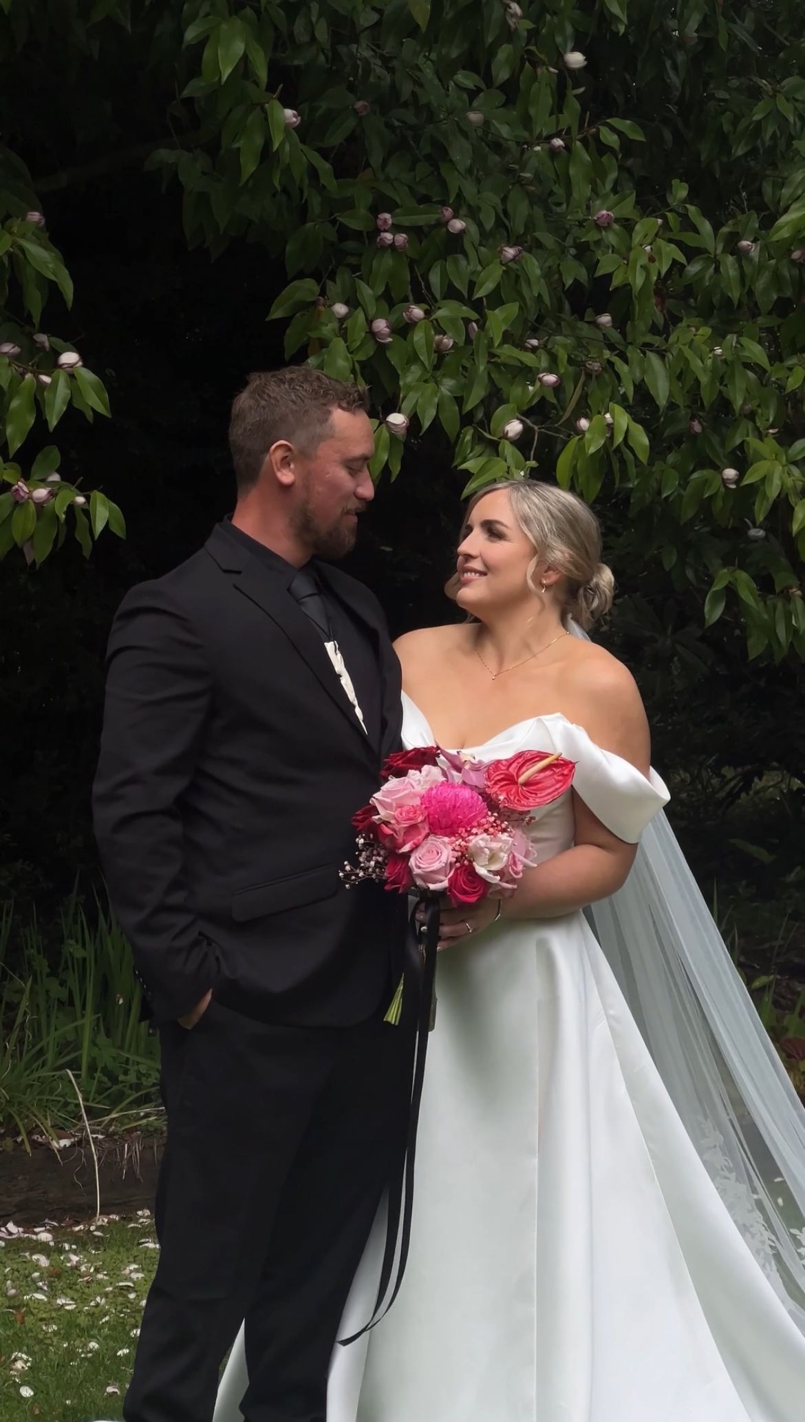 Bride in flowing white wedding dress with pink bouquet posing with groom in garden setting.