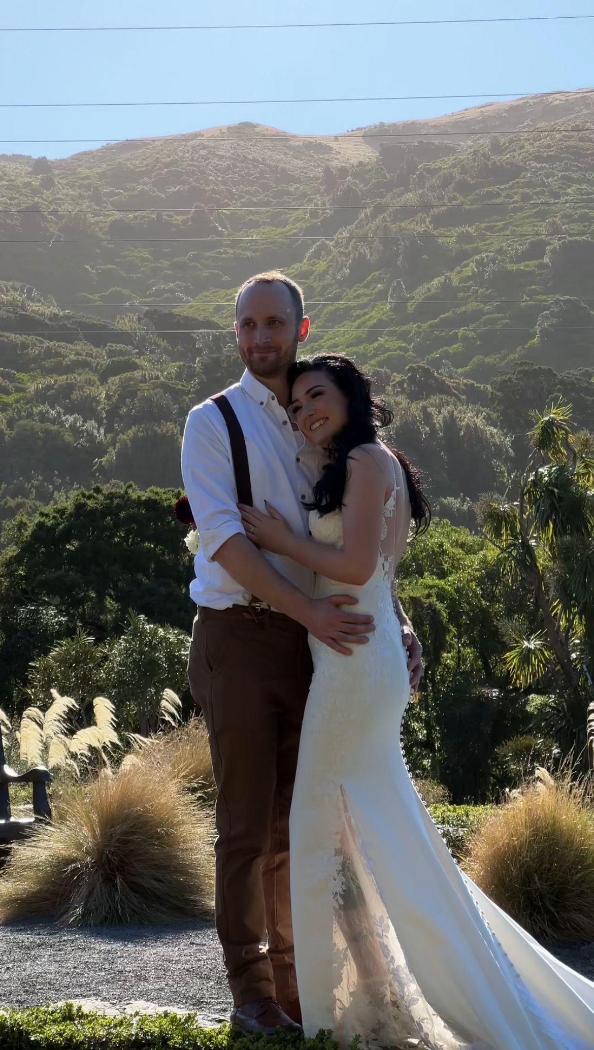 A couple embraces on a scenic hillside during their wedding celebration with mountains and natural landscape in the background.