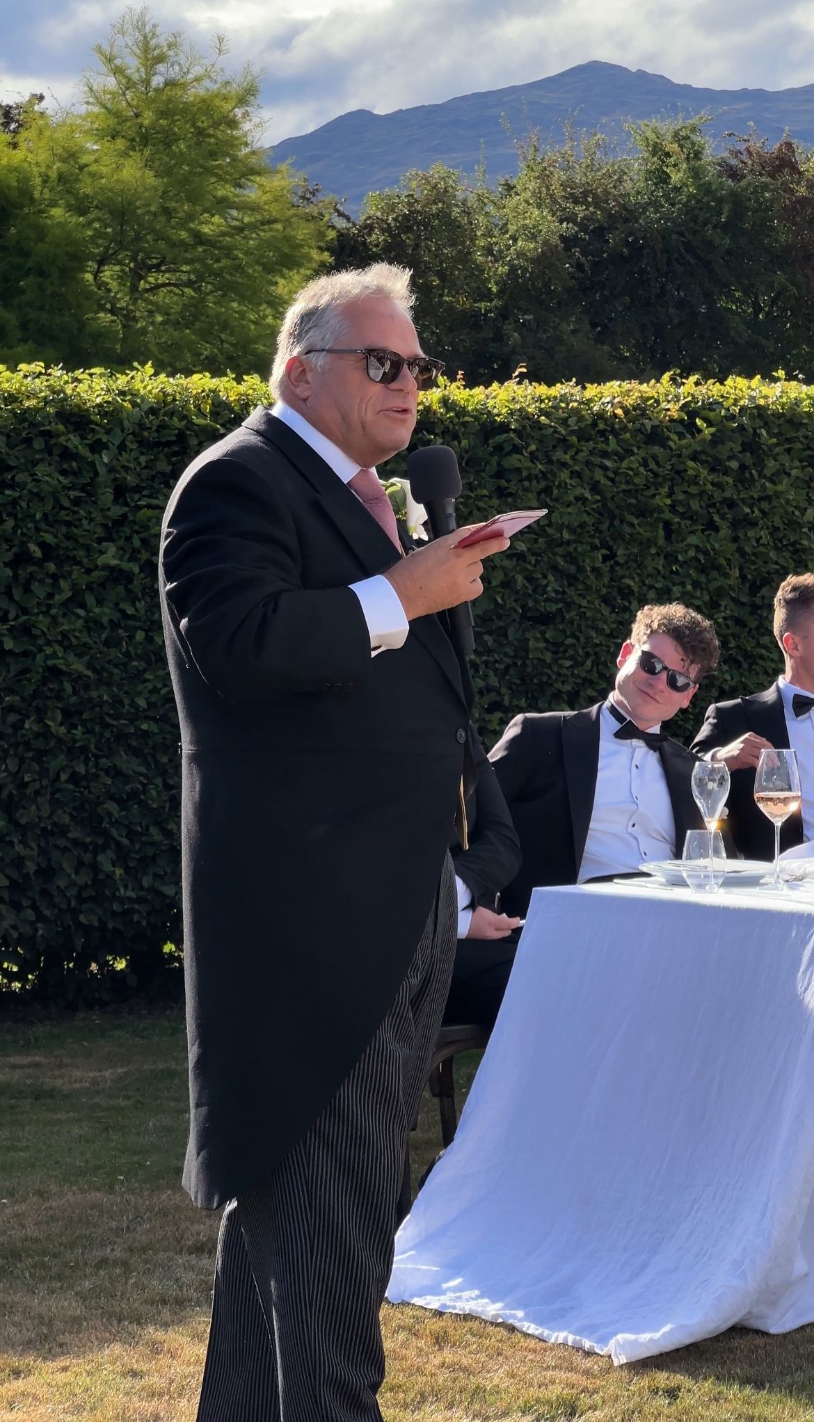 A series of photos showing a person in a black suit speaking at an outdoor event with white-clothed tables and mountain views.