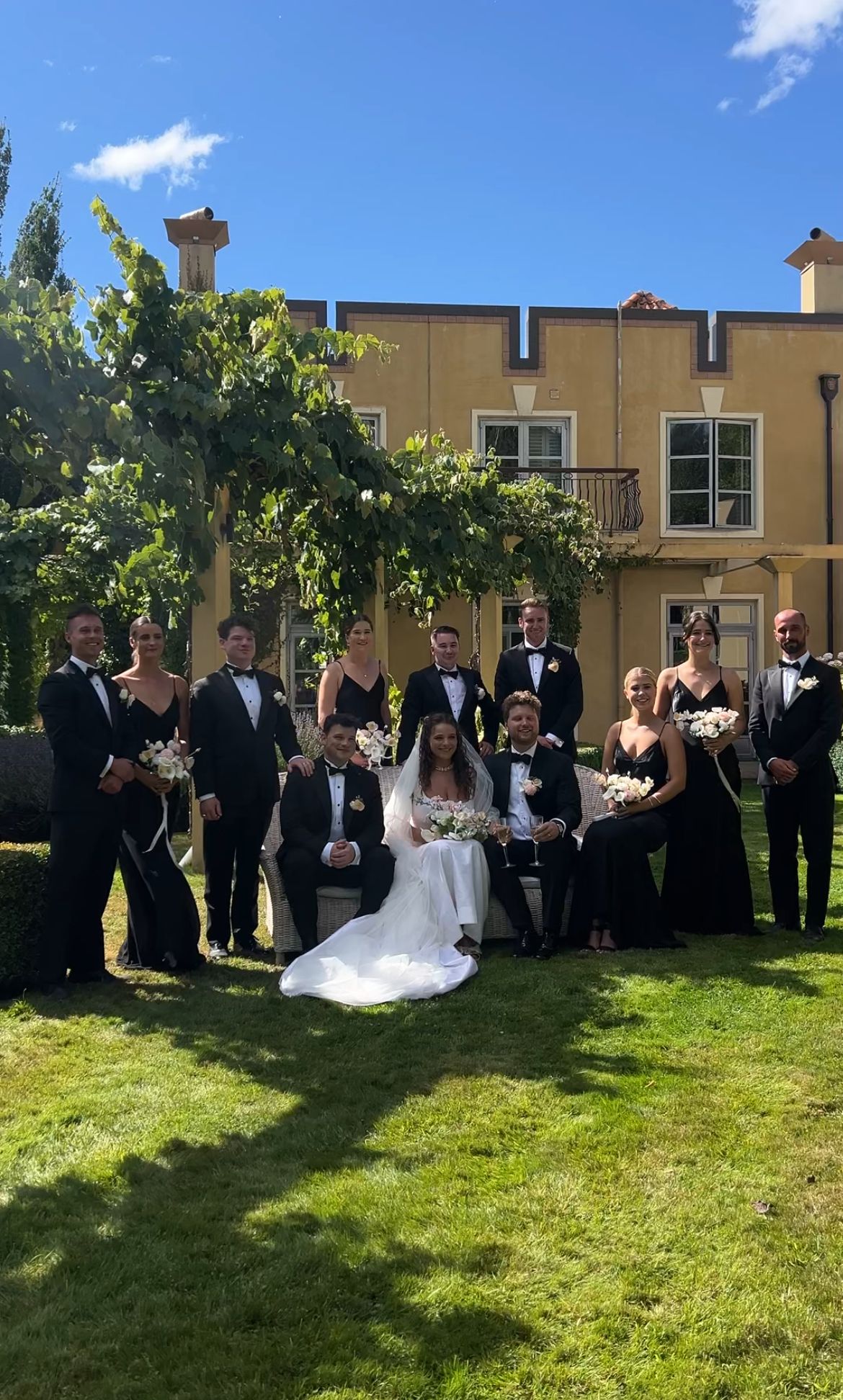 Wedding party in formal attire poses for photos on a sunny day in a grassy outdoor setting with a beige building backdrop.
