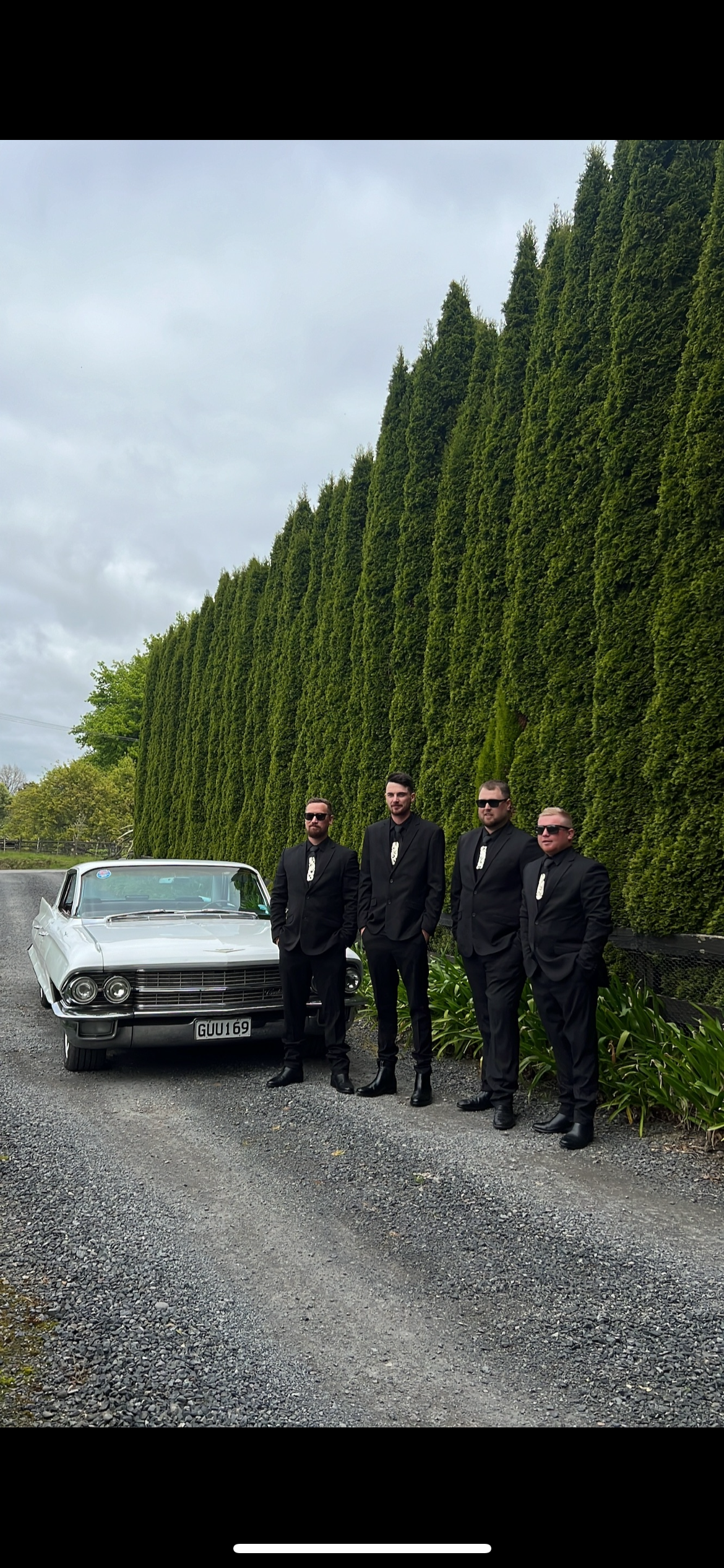 A classic white Cadillac parked alongside tall cypress trees with people in black suits standing nearby.