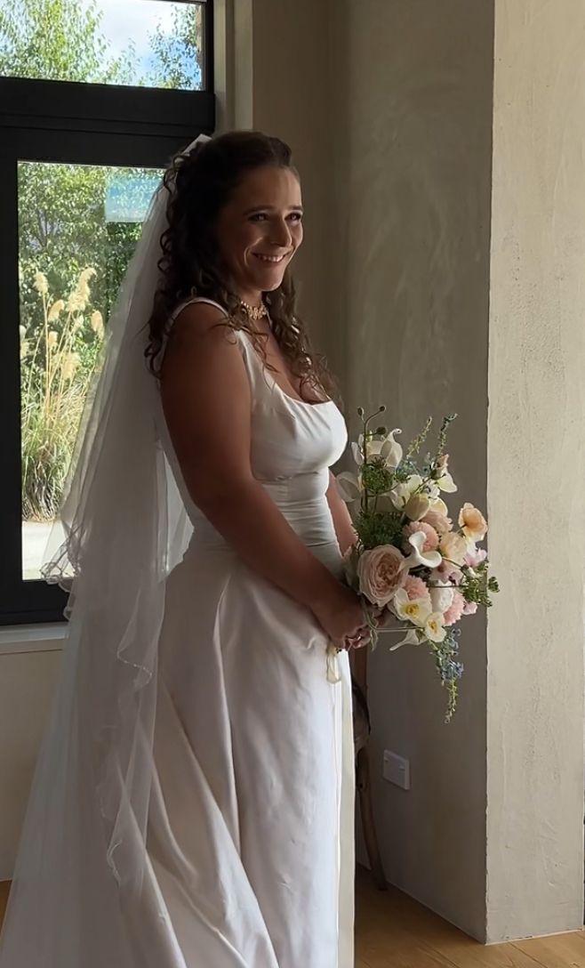 A bride in a flowing white dress holds a romantic bouquet of blush roses and delicate flowers in a sunlit room.
