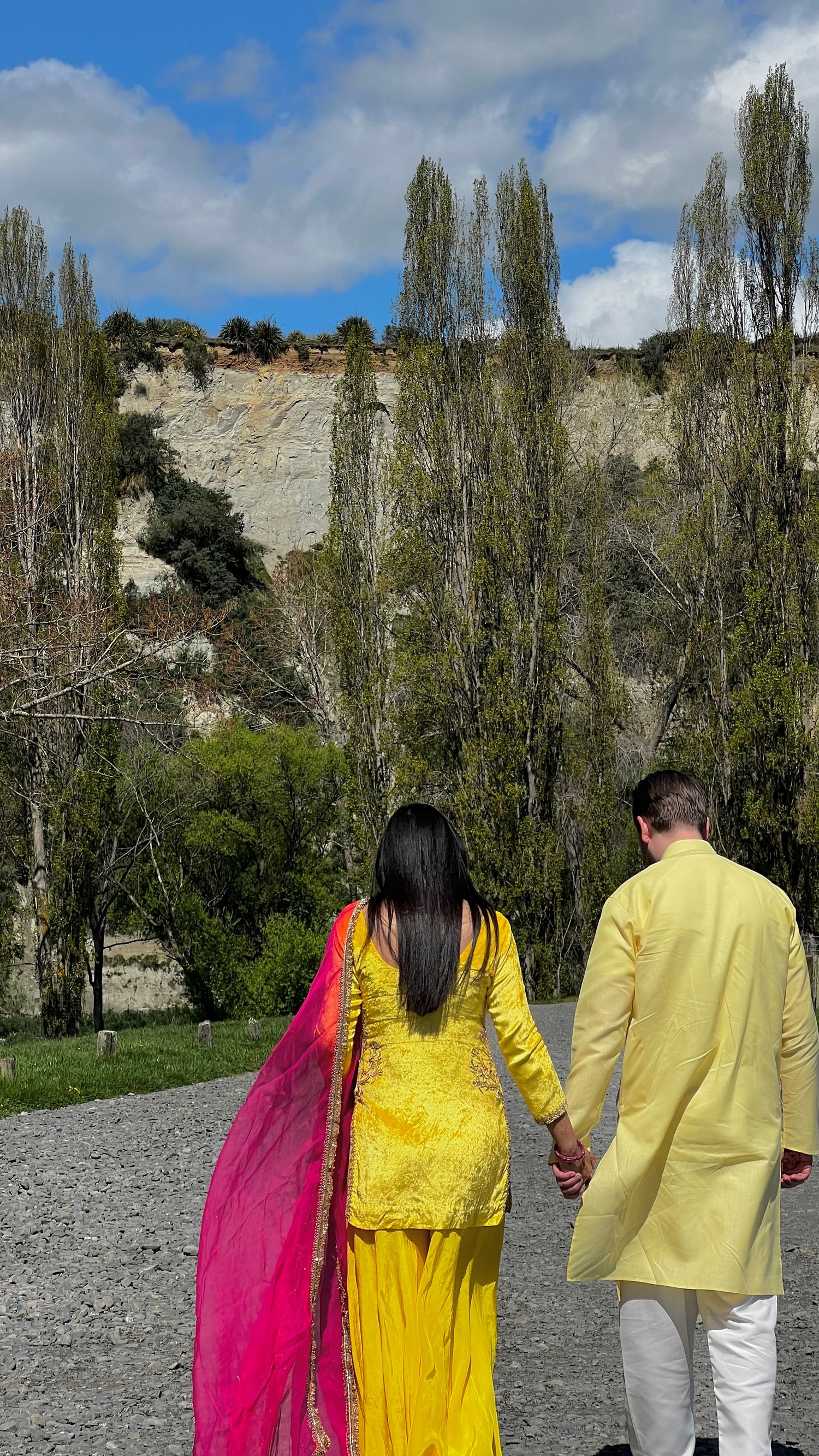 Couple in traditional indian yellow and pink attire walking along a path with tall poplar trees.