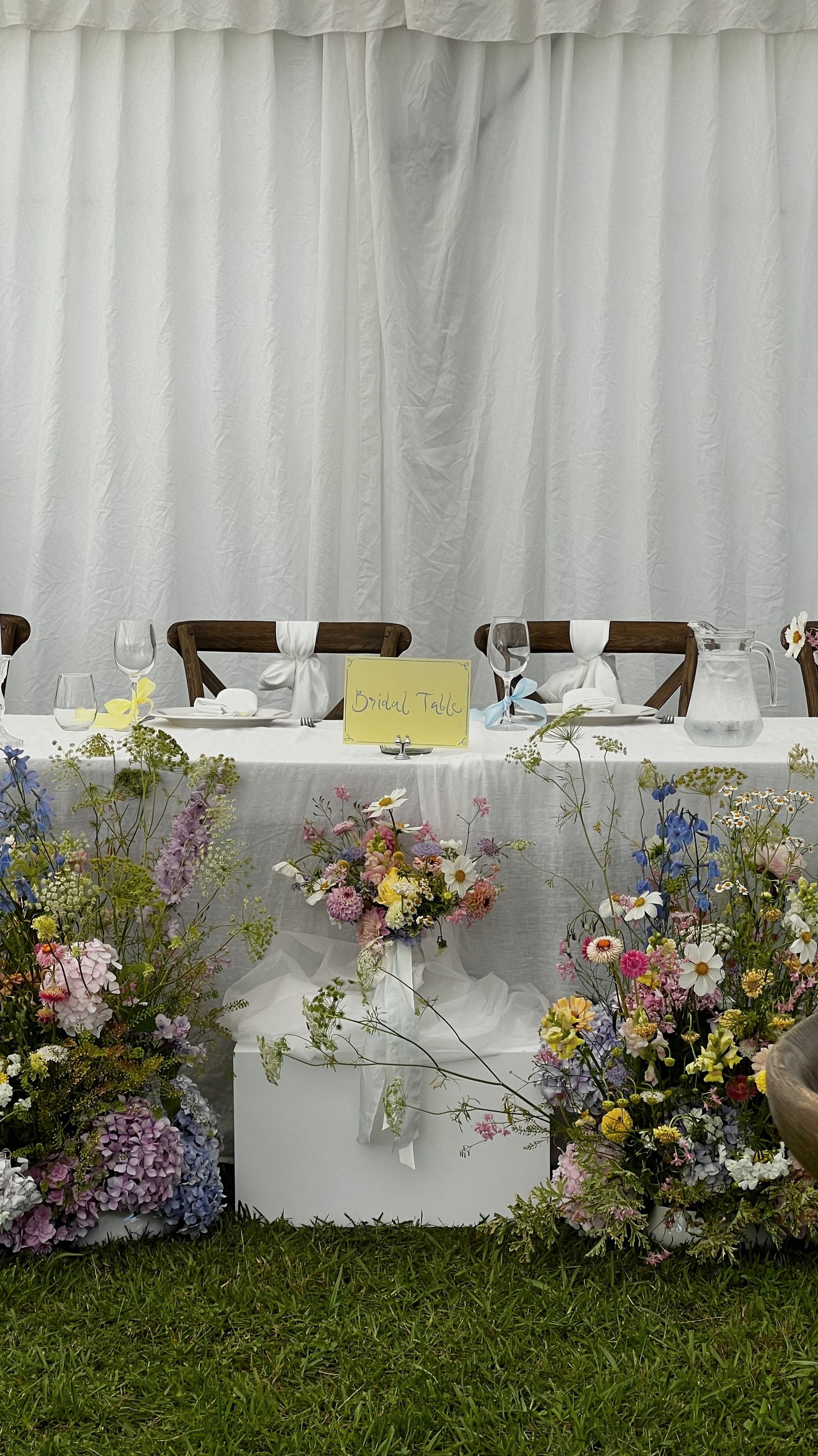 Wildflower arrangements line a white tent wall at an outdoor wedding reception.